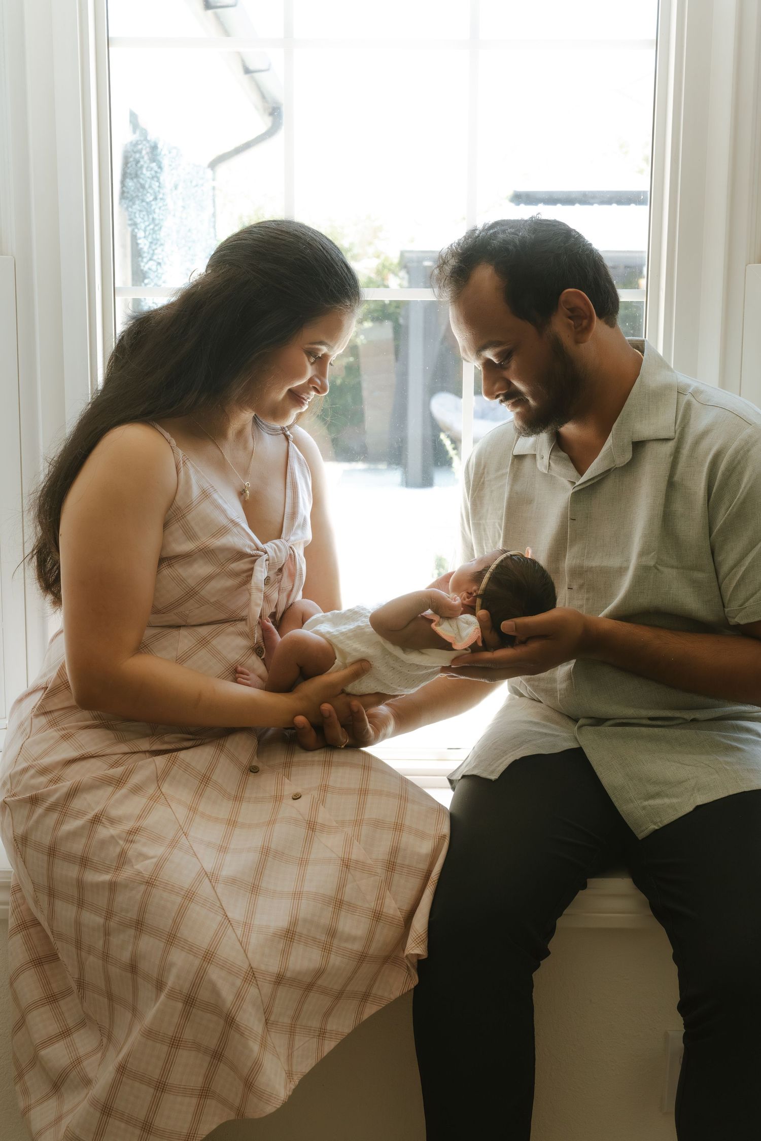 A couple sits by a bright window, lovingly gazing at their newborn baby in a warm, intimate family portrait.