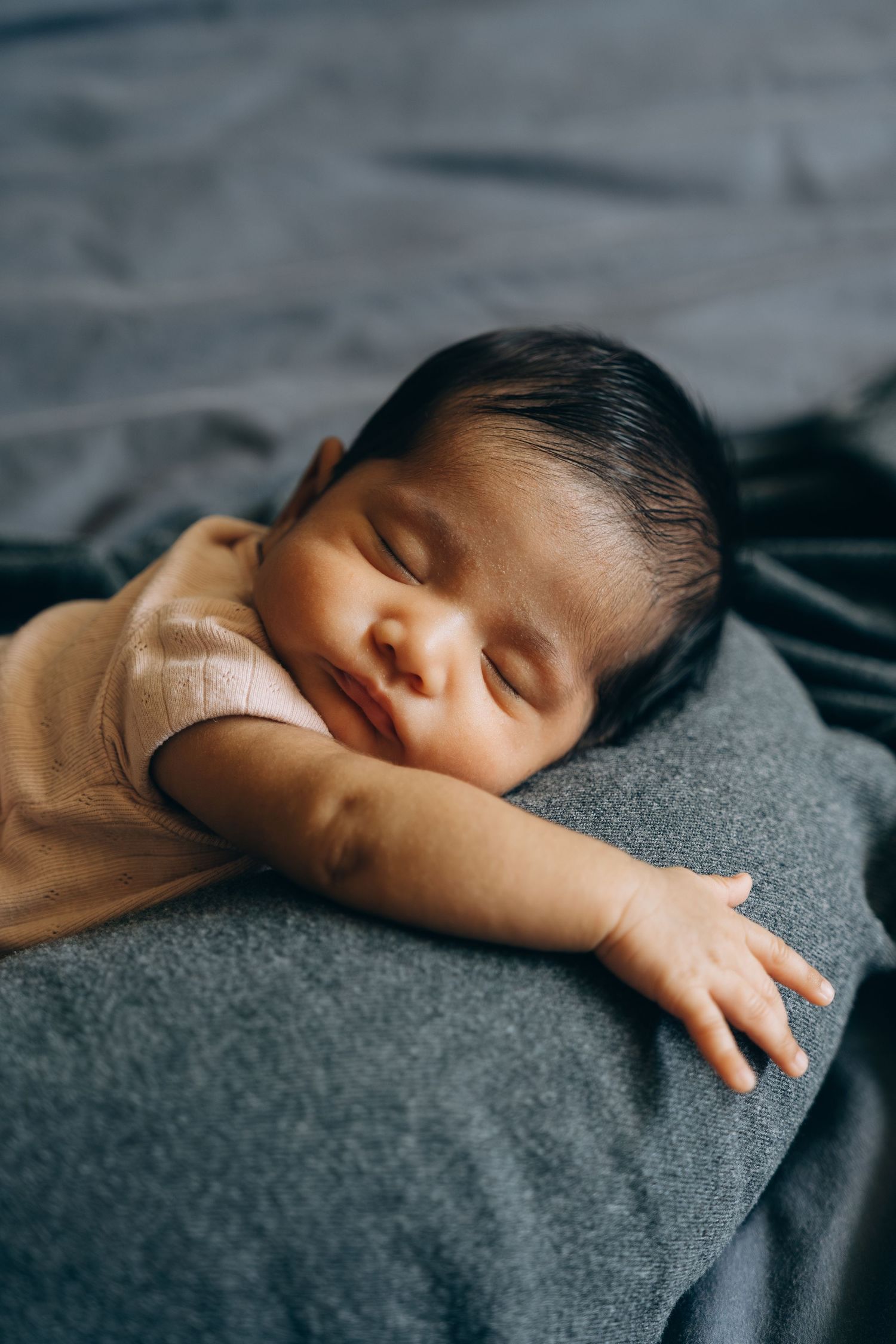 Peaceful newborn baby sleeping on a gray blanket, wearing a pink onesie, with dark hair and a gentle smile.