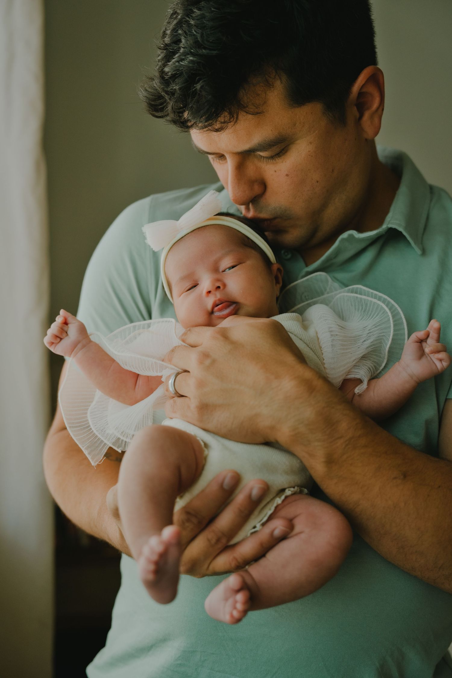 A father tenderly kisses his sleeping newborn baby dressed in white with a bow headband.