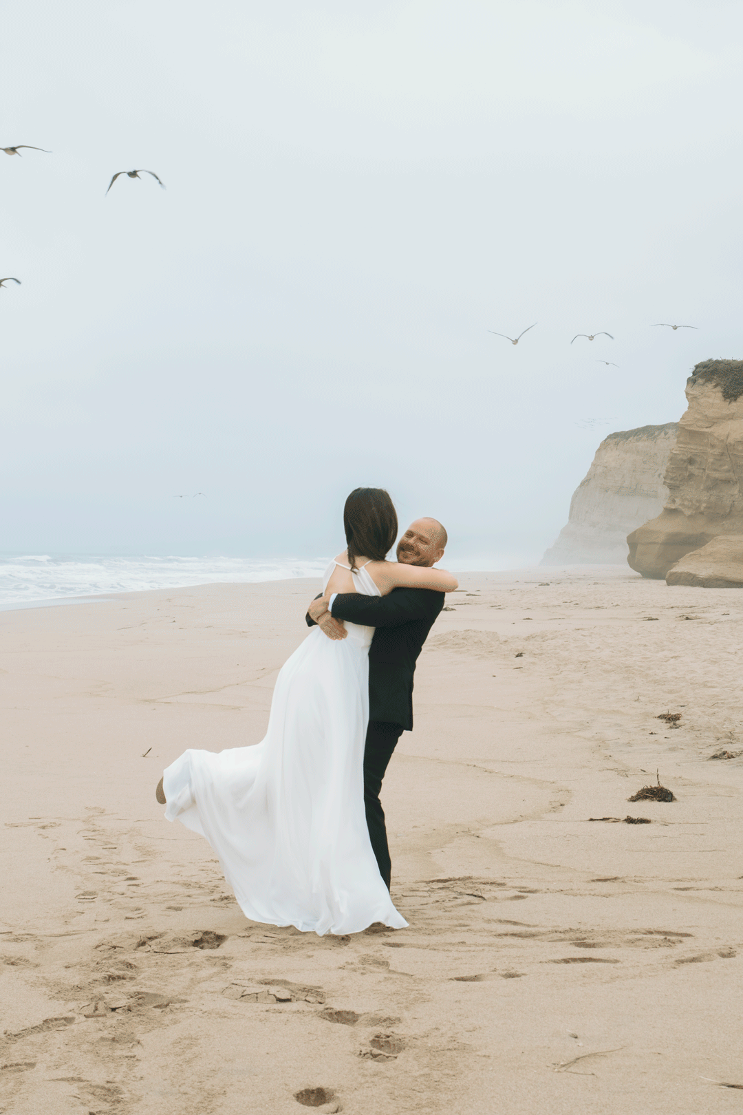 A couple embraces on a sandy beach, the bride in a flowing white gown, with seagulls and cliffs in the background.