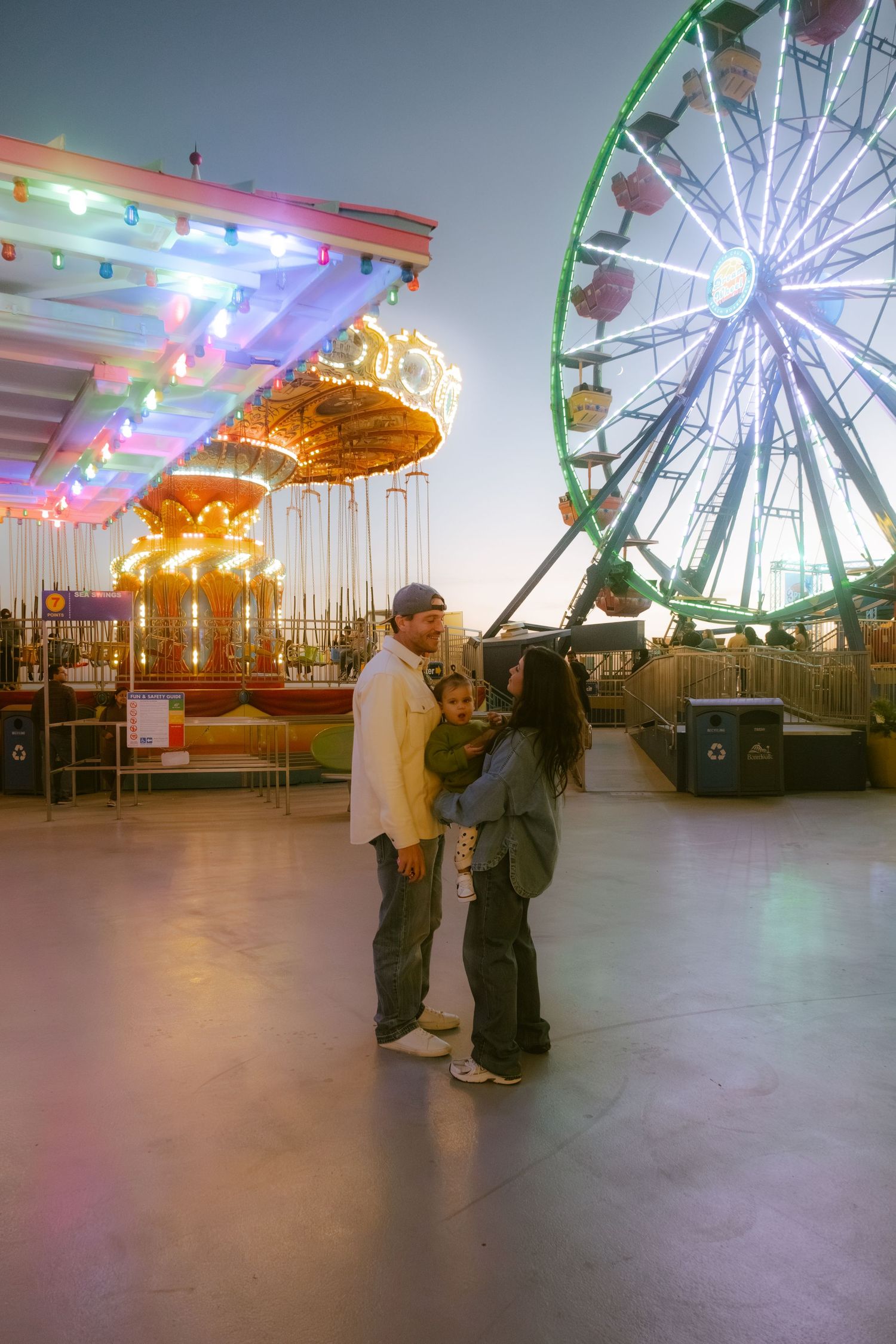 Couple with child at amusement park at dusk, with lit Ferris wheel and carousel in background.