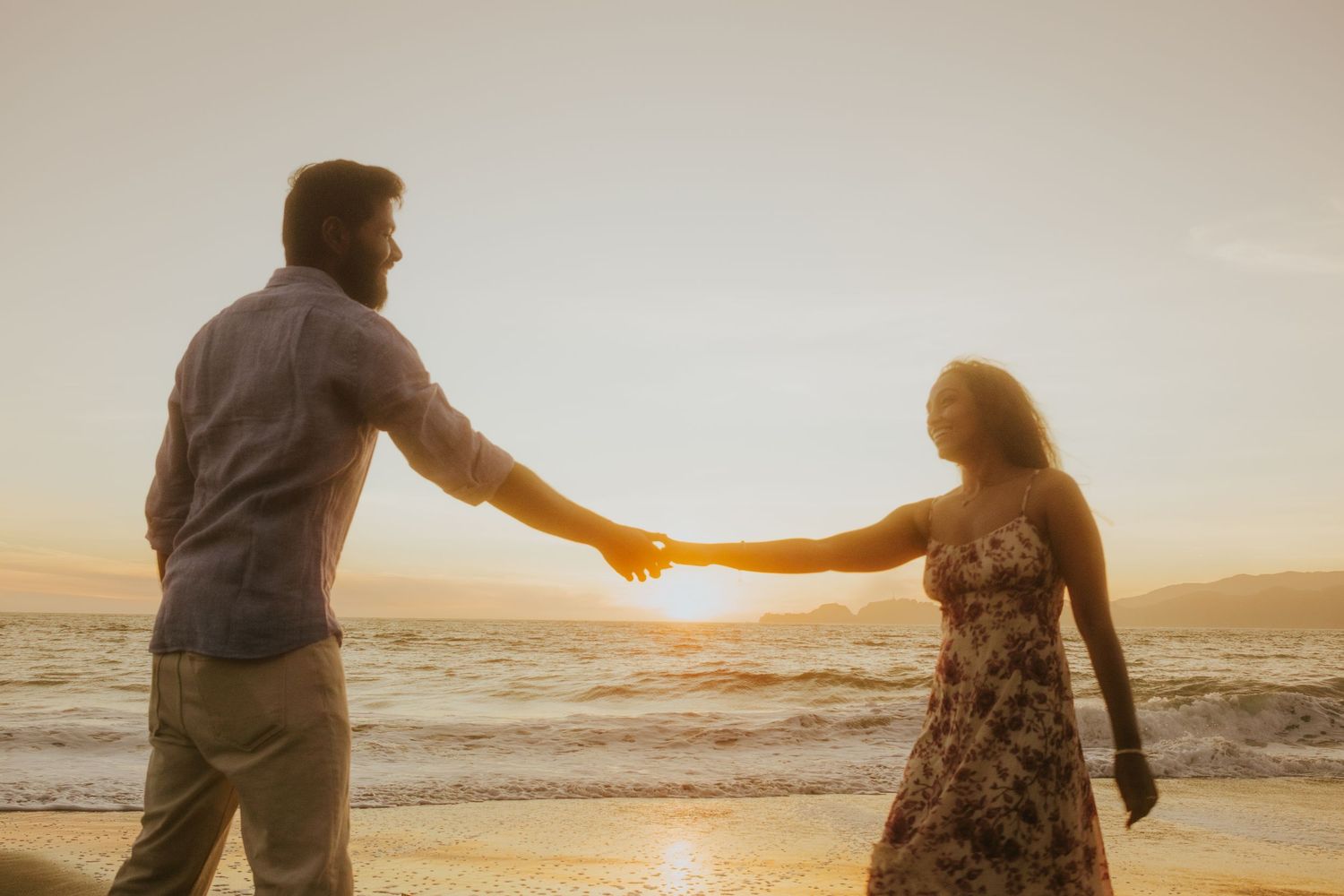 Romantic couple holding hands at sunset on a beach with golden light reflecting on the ocean waves.