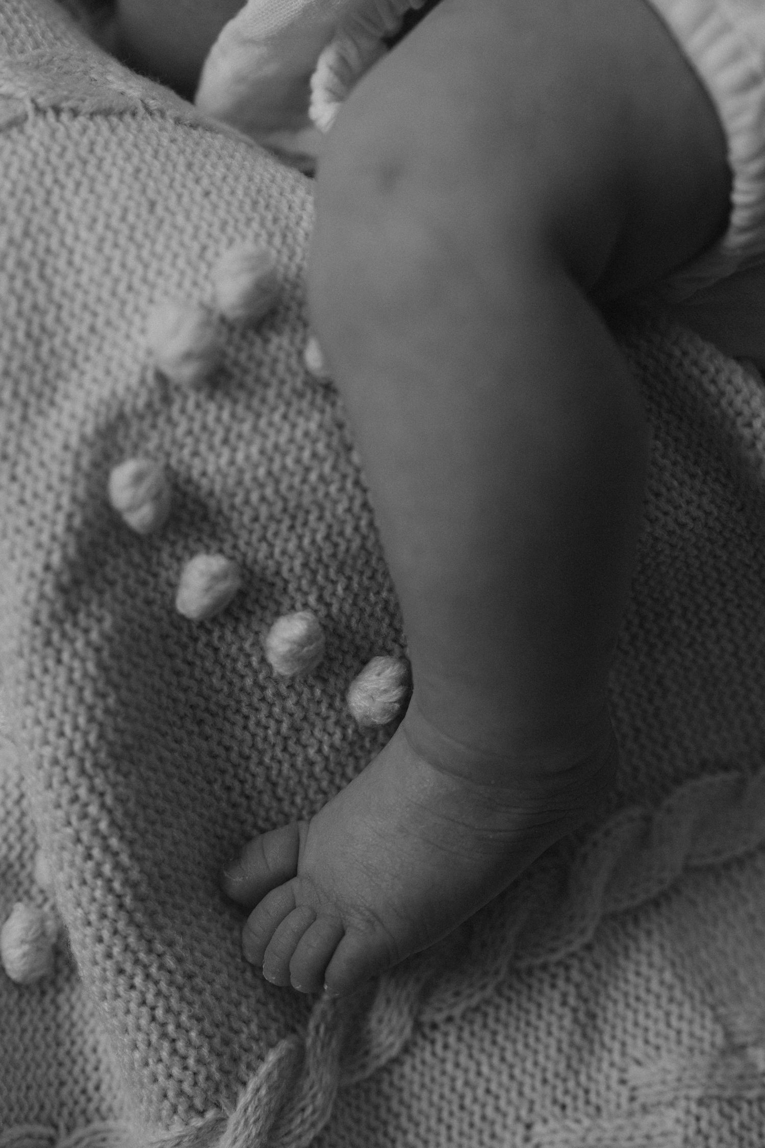 Black and white close-up photo of a newborn baby's tiny feet and legs resting on a soft knitted blanket.