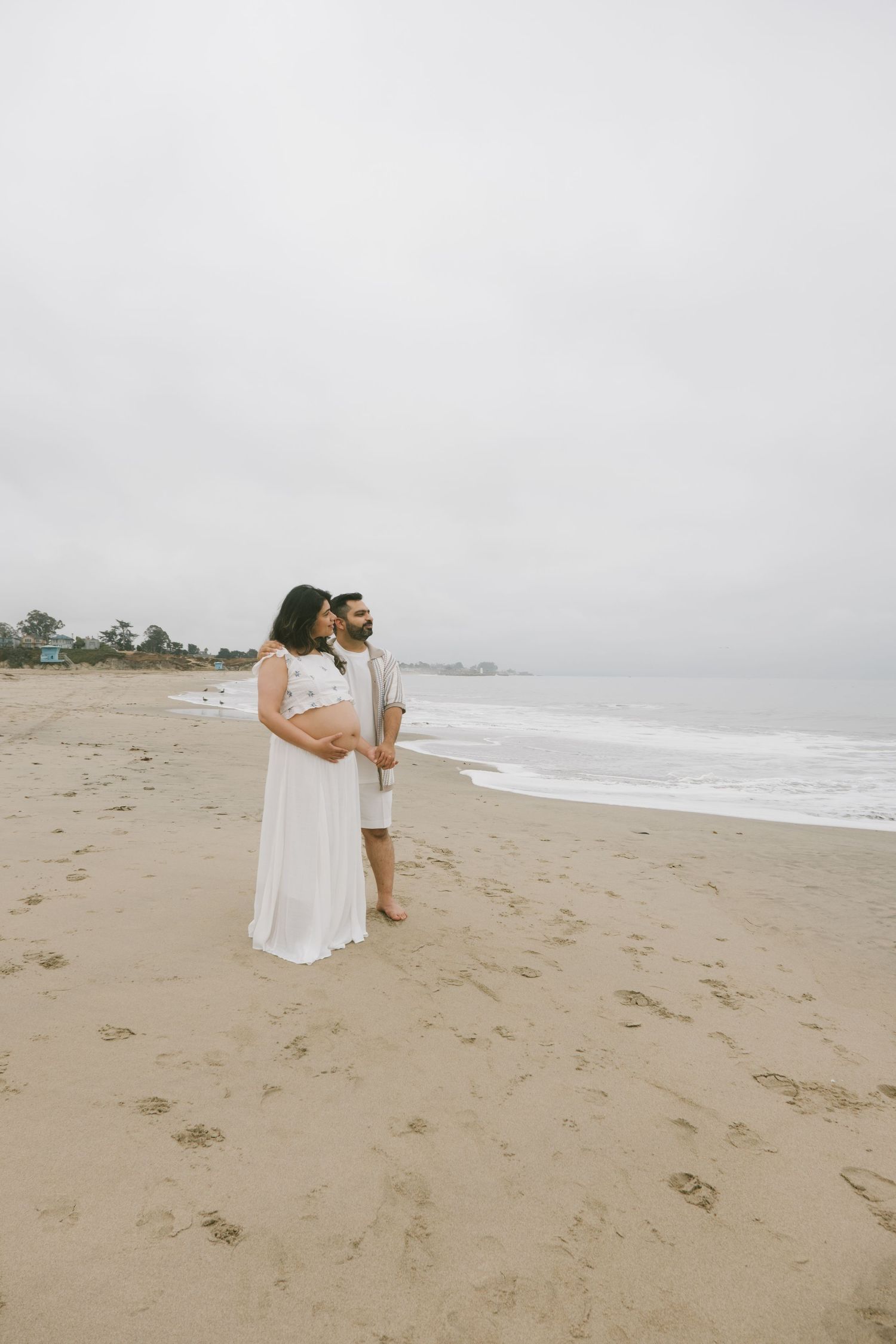 A couple in white and beige outfits poses together on a misty beach.