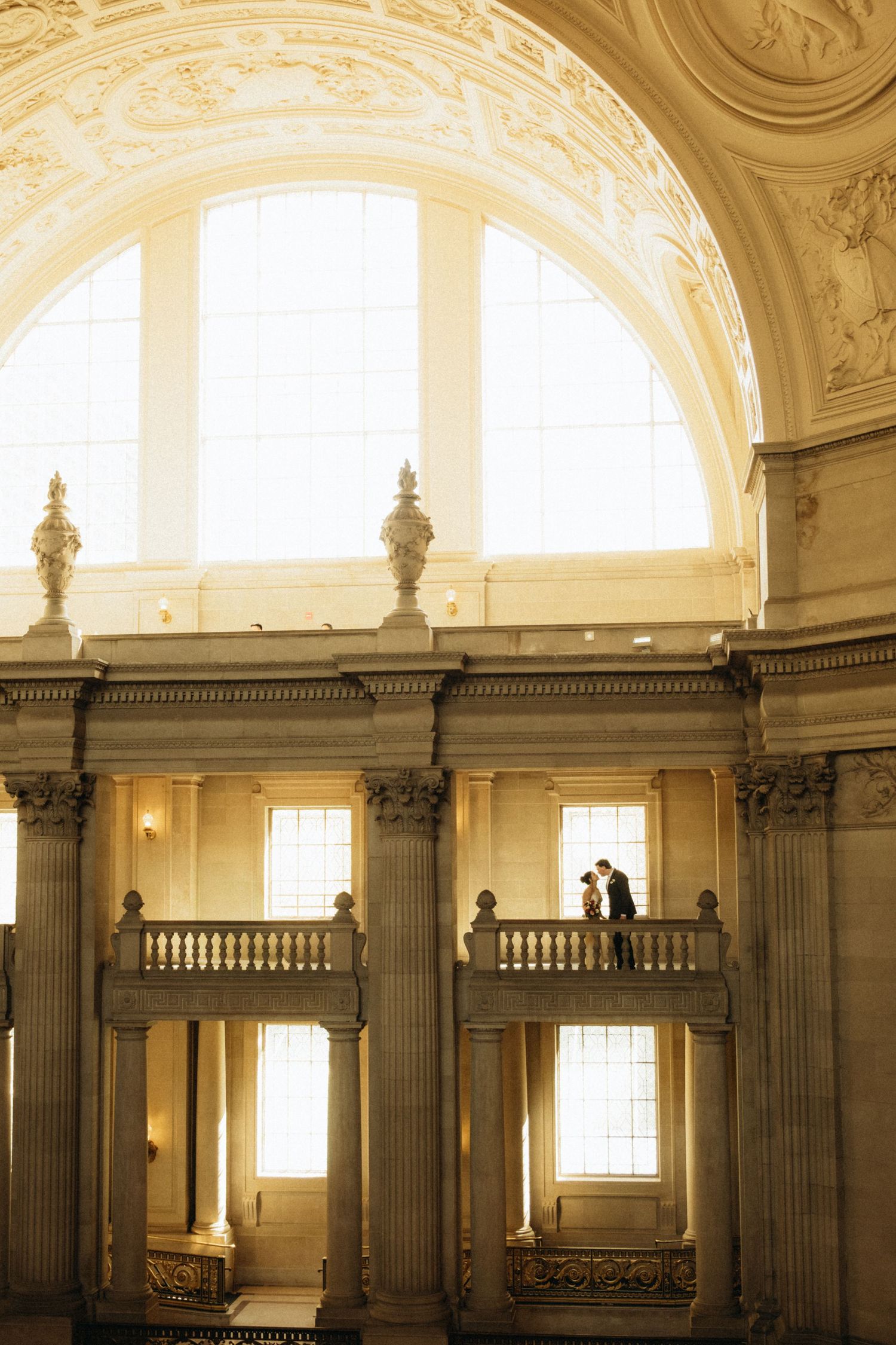 Couple sharing a romantic moment on a grand balcony inside a ornate neoclassical building with arched windows.