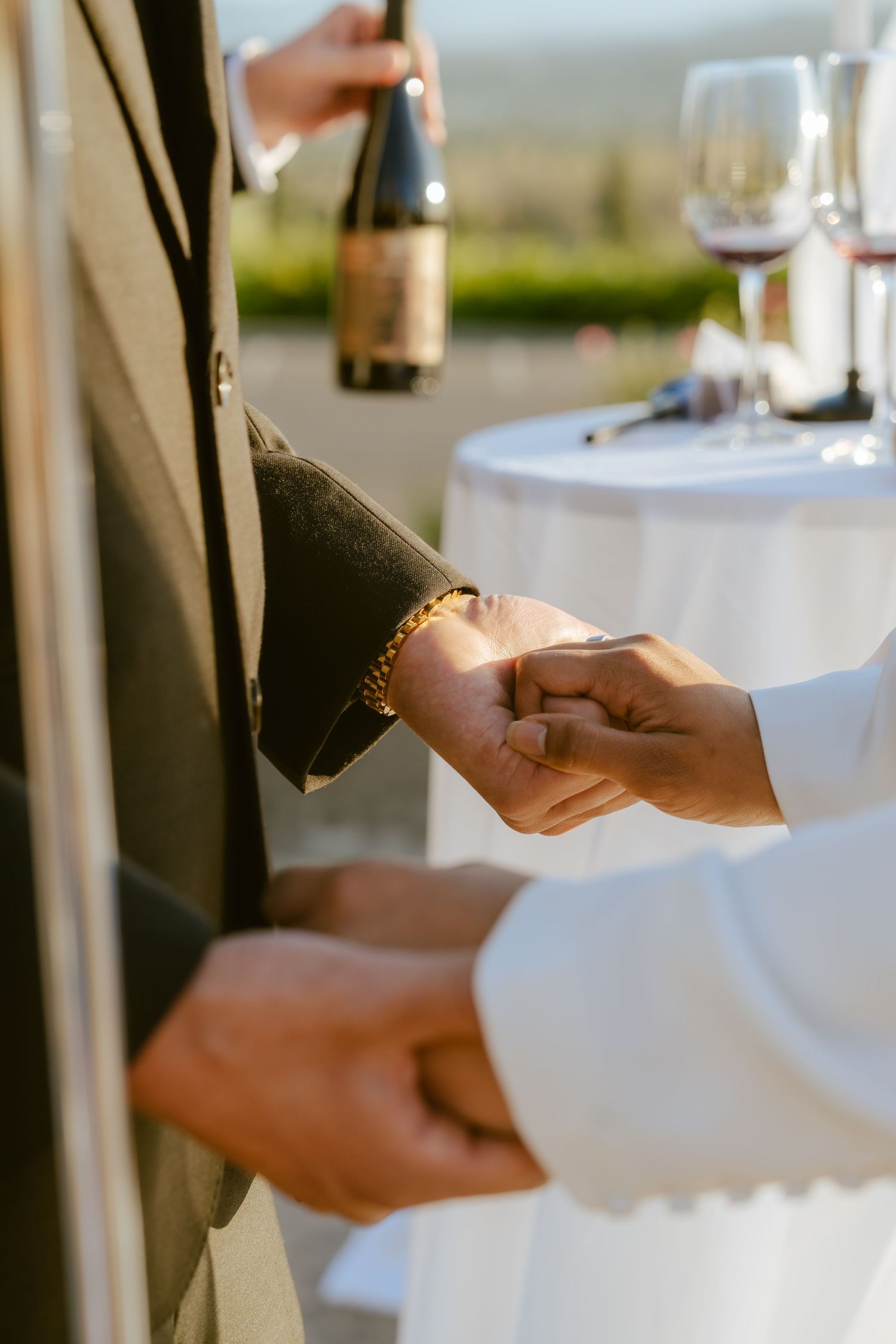 A person wearing a gold bracelet has their hand held at an outdoor wedding reception with wine bottles in background.