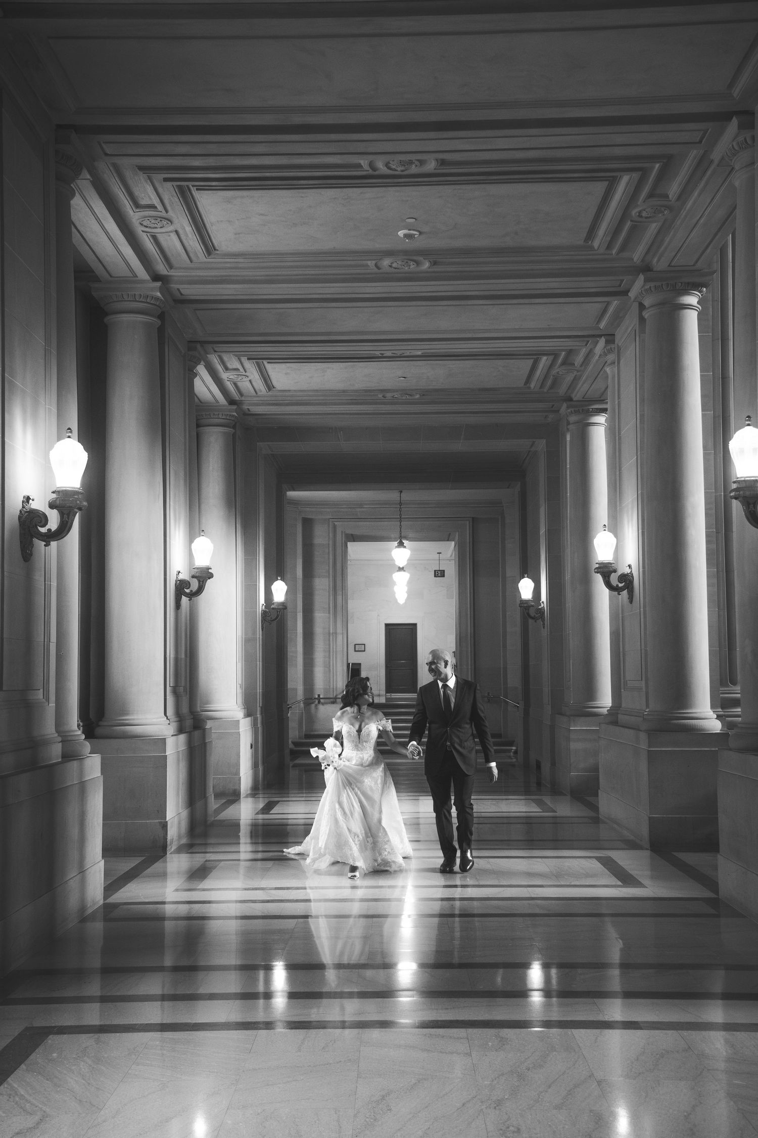 Black and white wedding photo in a dramatic marble hallway with classical lighting fixtures.