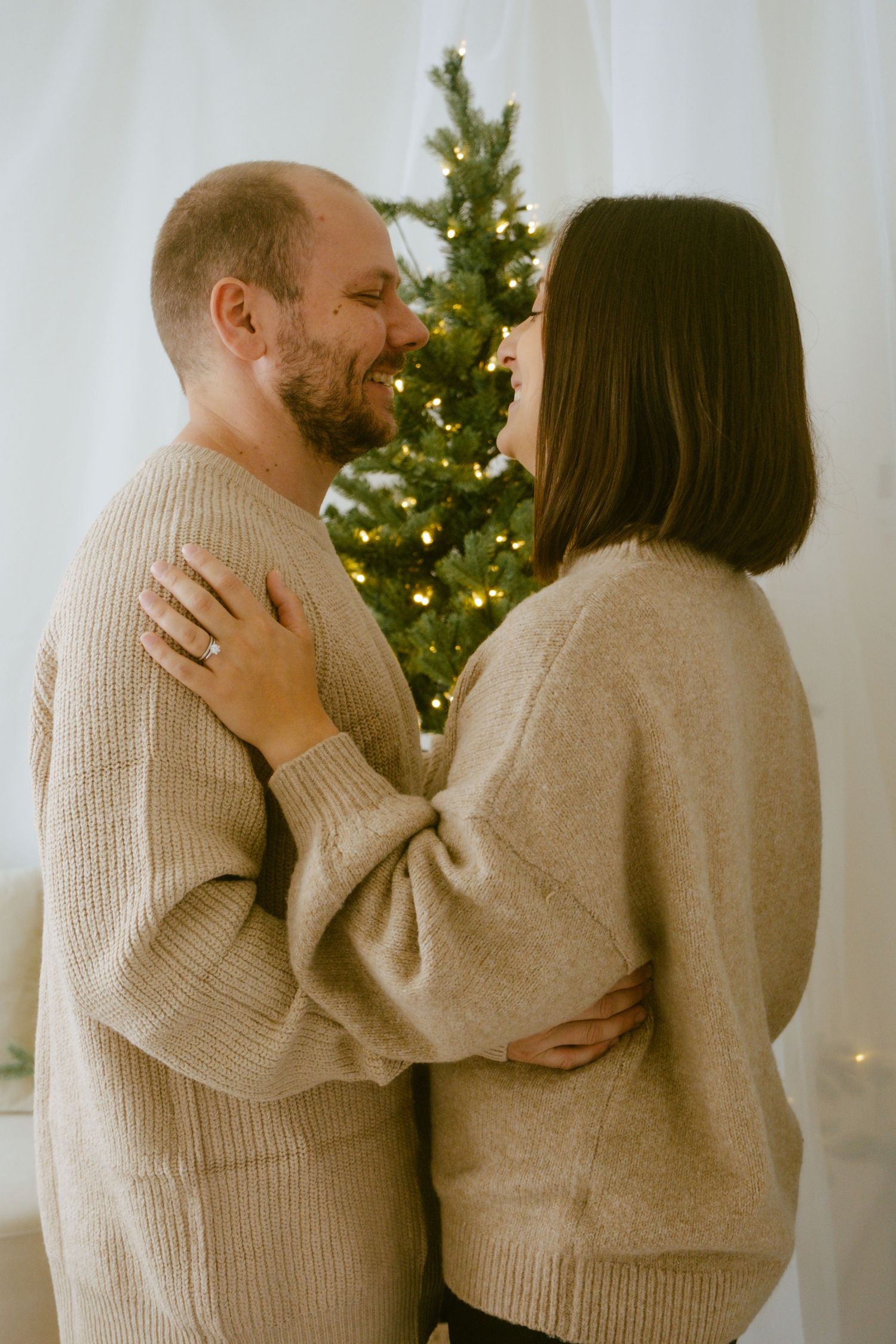 Couple in matching beige sweaters embrace in front of a lit Christmas tree in a cozy indoor setting.