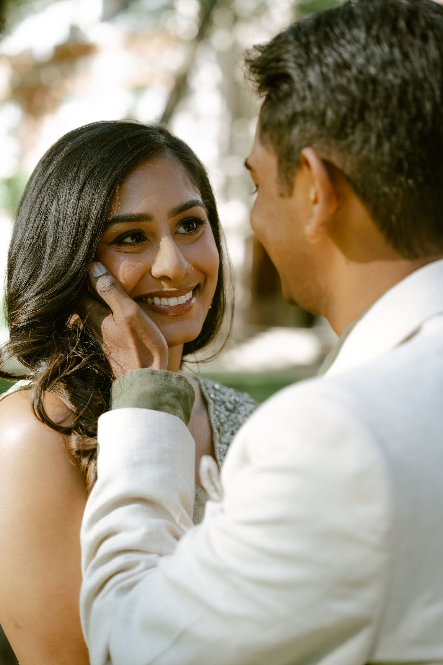 A groom in a white suit tenderly touches his smiling bride's face during an outdoor wedding ceremony.