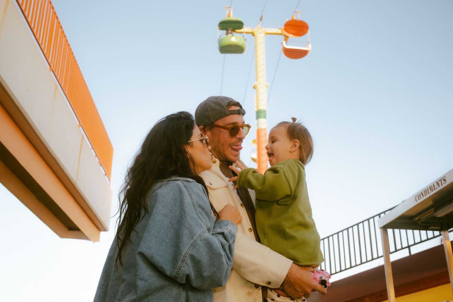 A couple holding a baby outdoors near a colorful amusement ride under a clear blue sky.