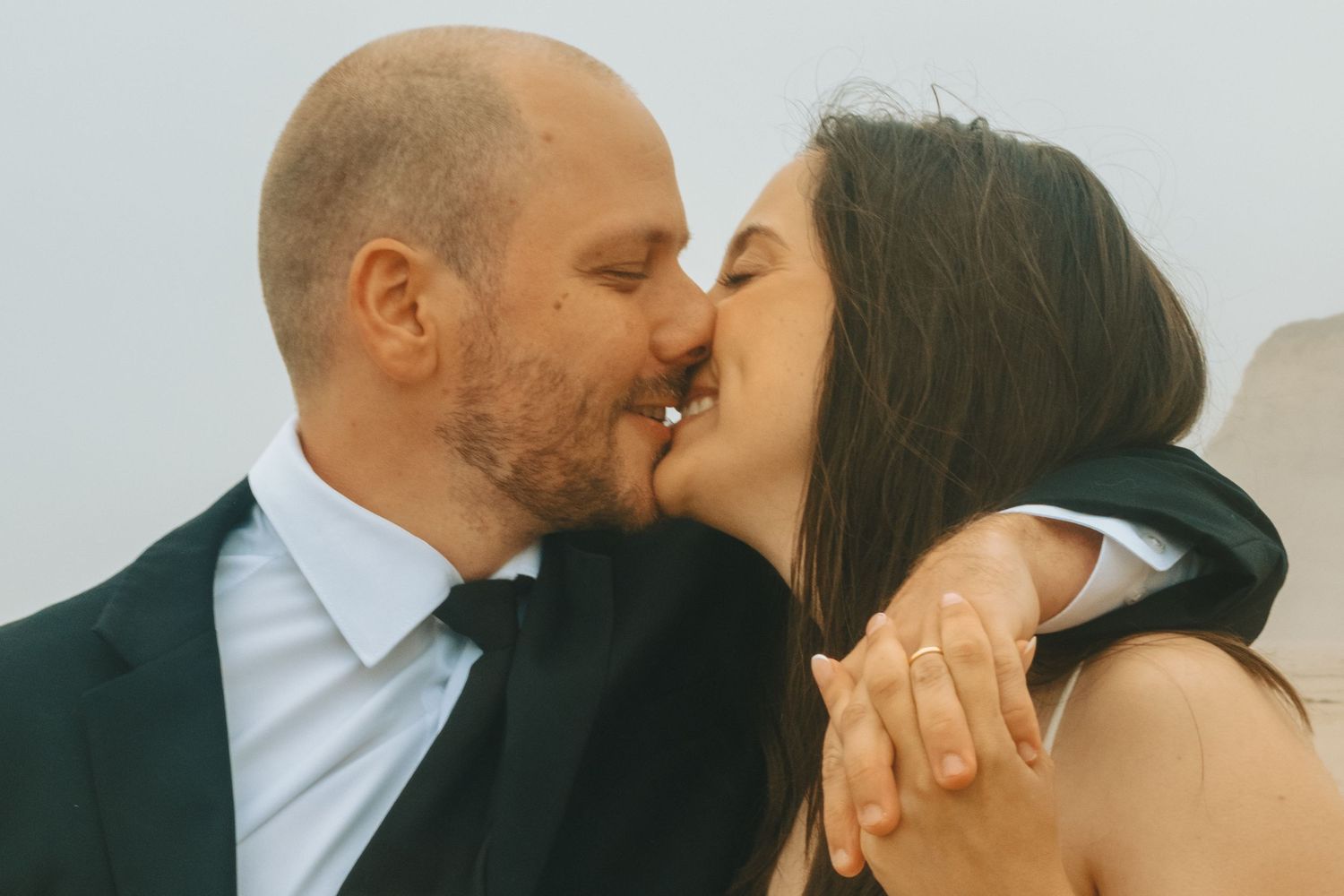 A couple shares a romantic kiss, the man in a suit and woman in a white dress, likely at their wedding.