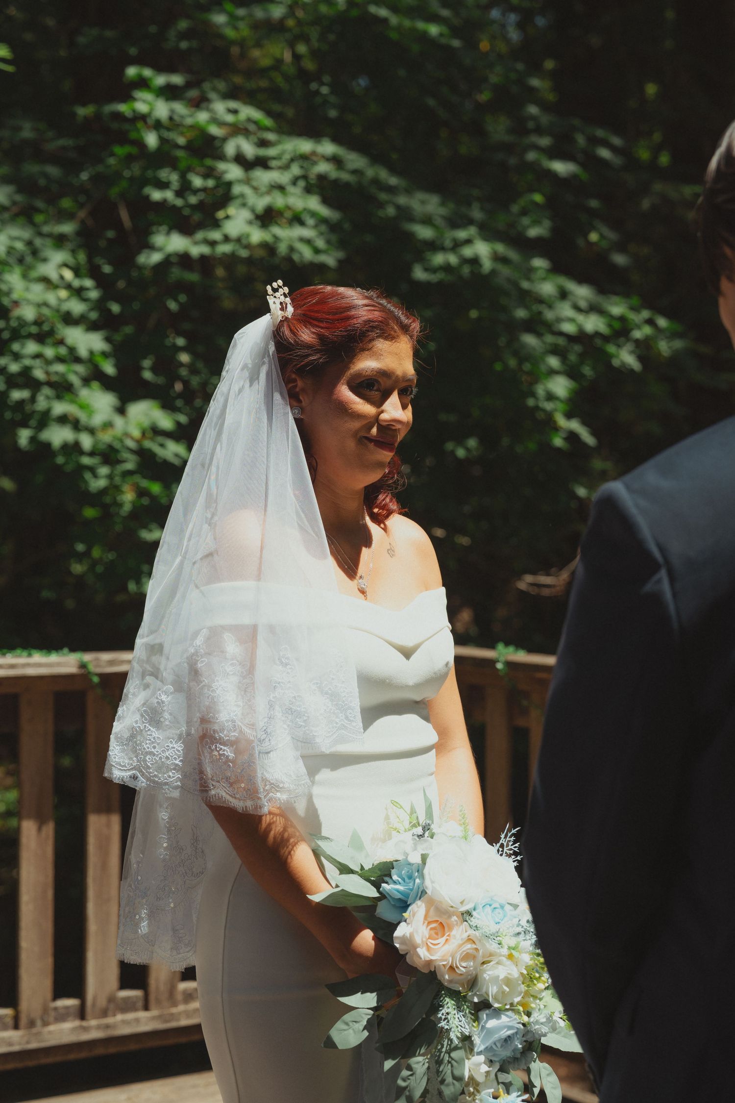 Bride in white gown and lace veil holds pastel bouquet outdoors at wedding ceremony.