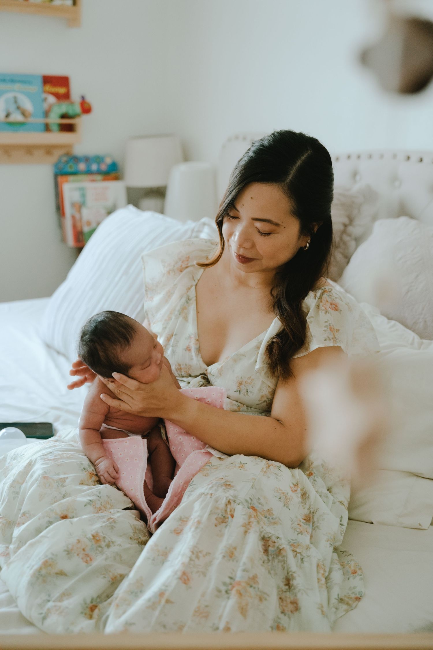 A mother in a floral dress tenderly holds her newborn baby while sitting on a white bed.