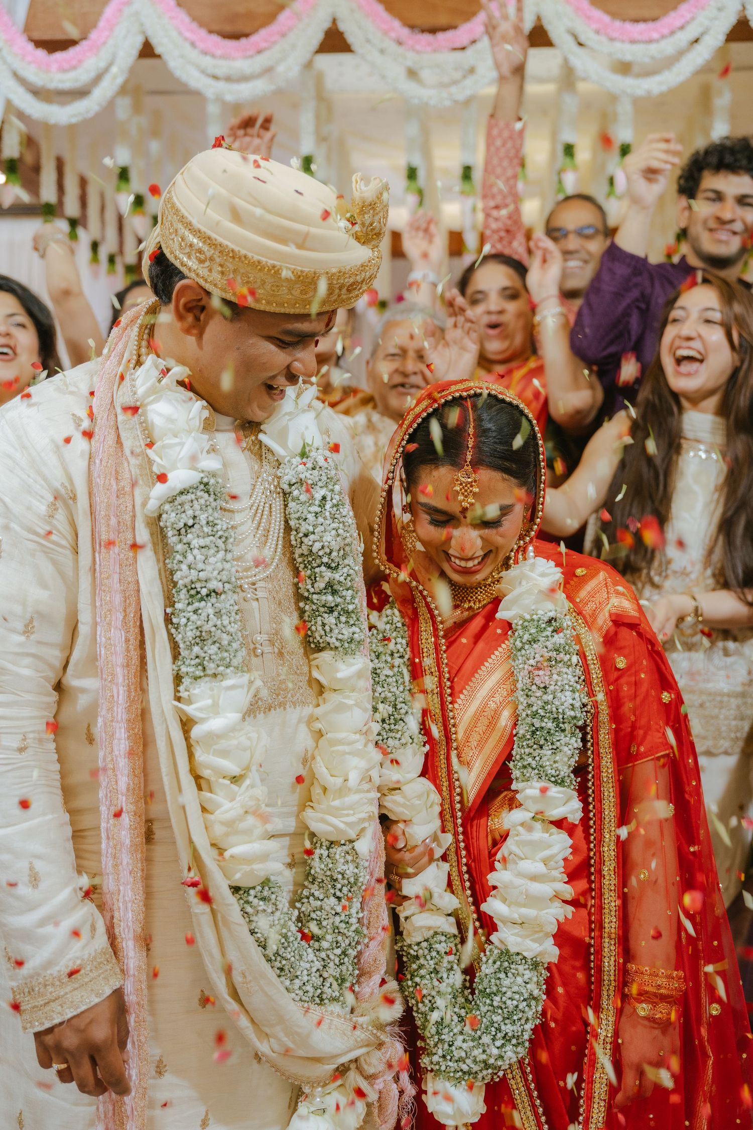Indian bride in red saree and groom in white sherwani exchange garlands at traditional Hindu wedding ceremony.