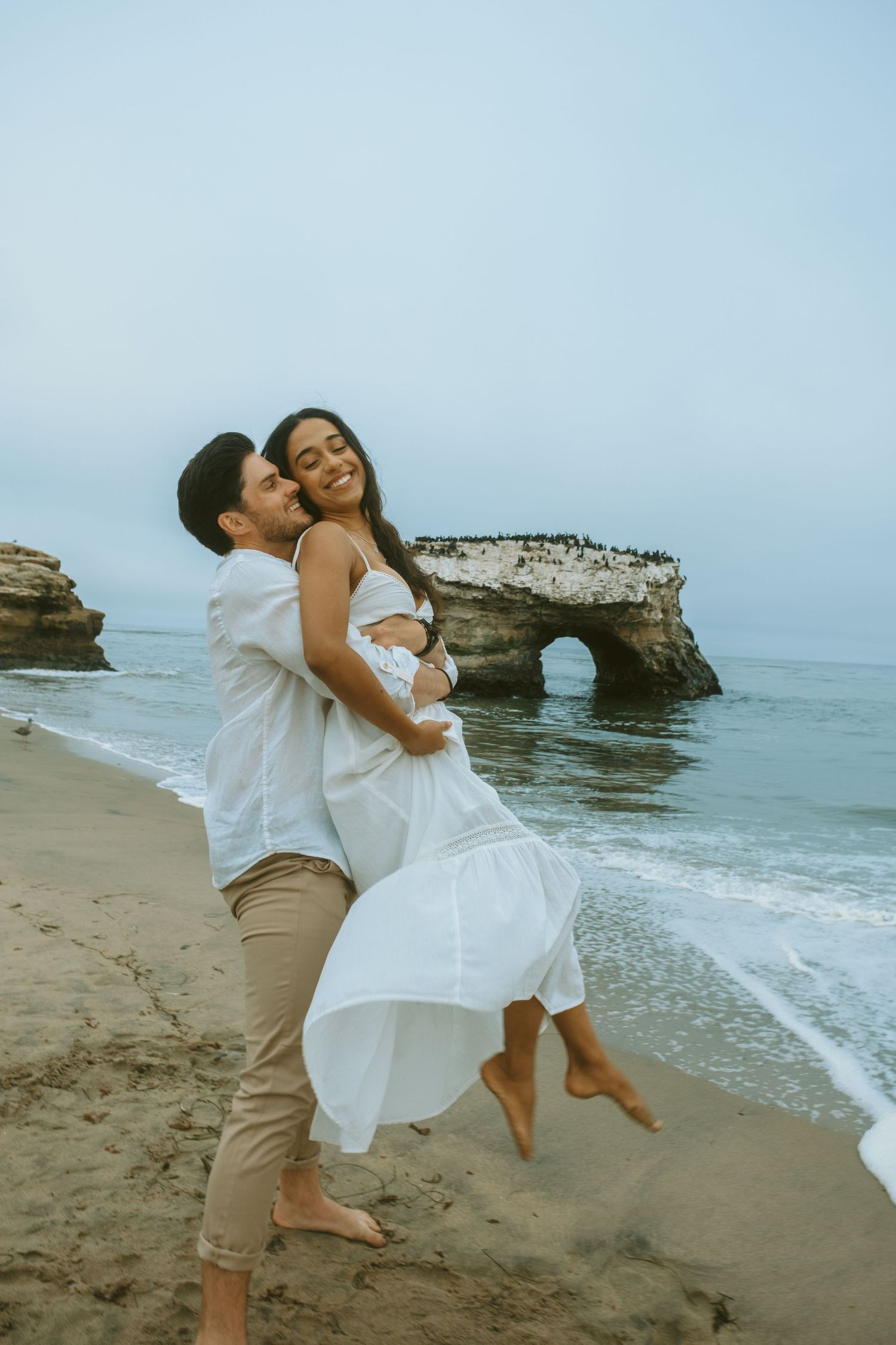 Playful couple in white outfits embracing on beach with natural rock formations.