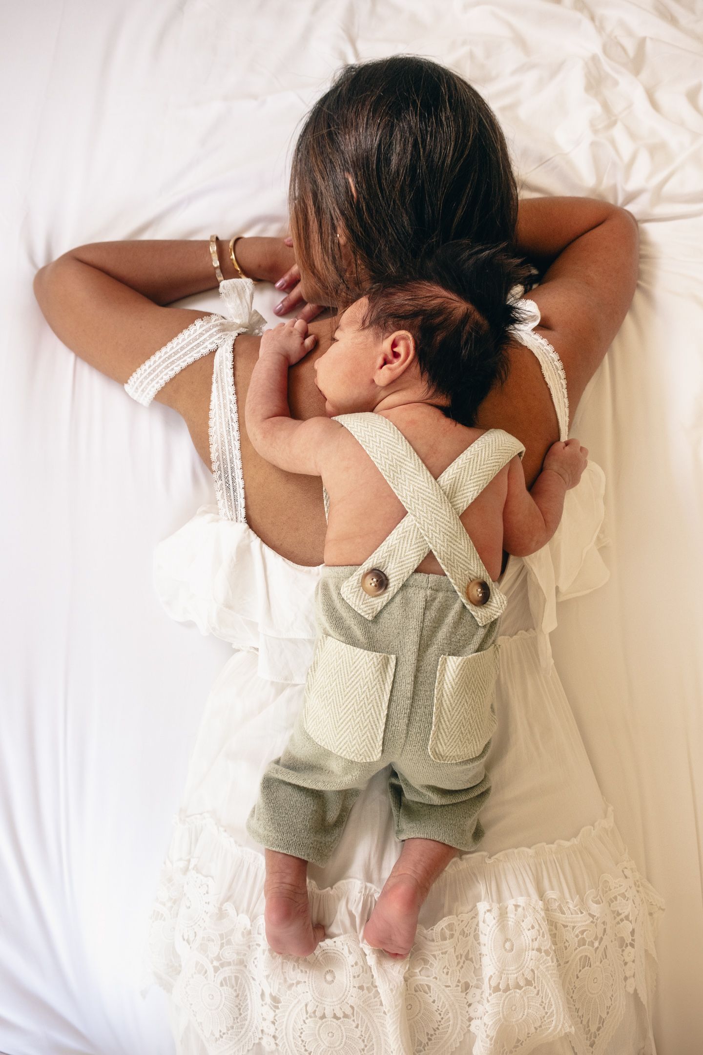 Baby wearing light green overalls lying on white lace bedding.
