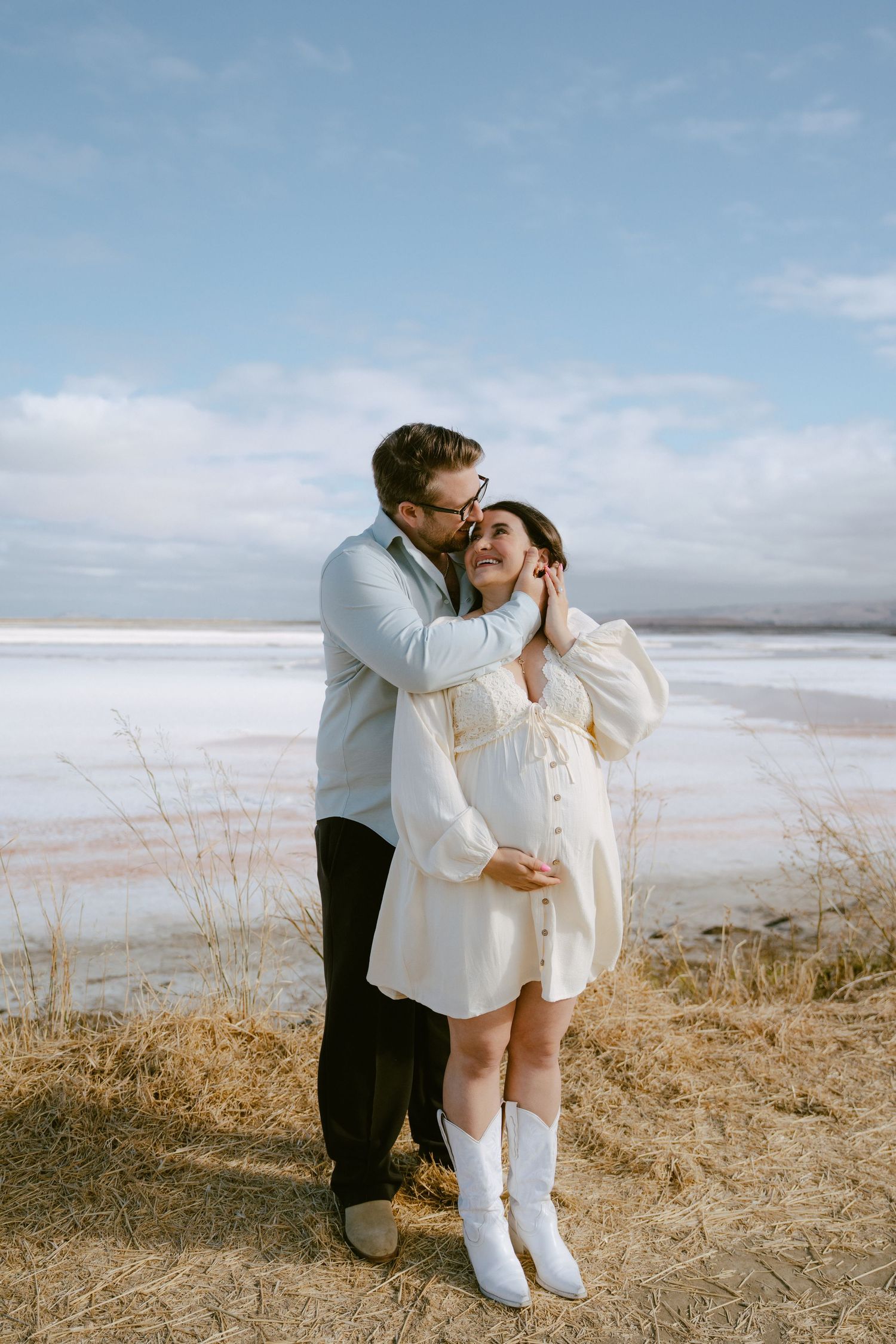 A couple embraces outdoors near a frozen lake, woman in cream dress and white boots, man kissing her cheek tenderly.