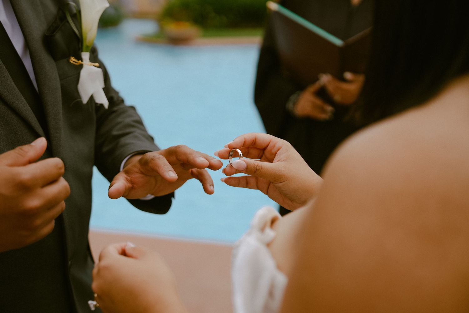 Bride placing wedding ring on groom's finger during outdoor ceremony near pool.