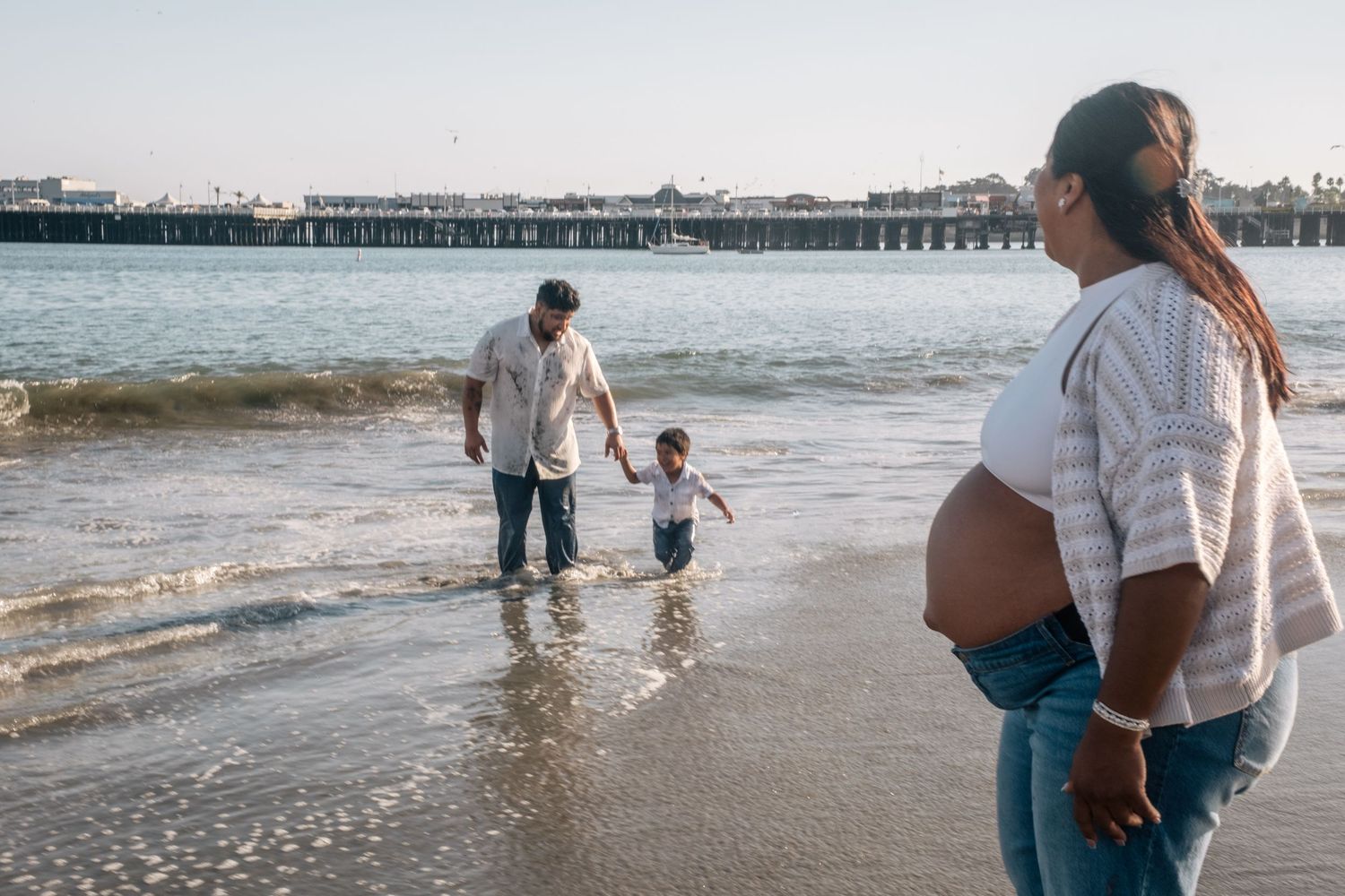 Pregnant woman watches husband and child play in ocean waves near a pier on a sunny beach day.