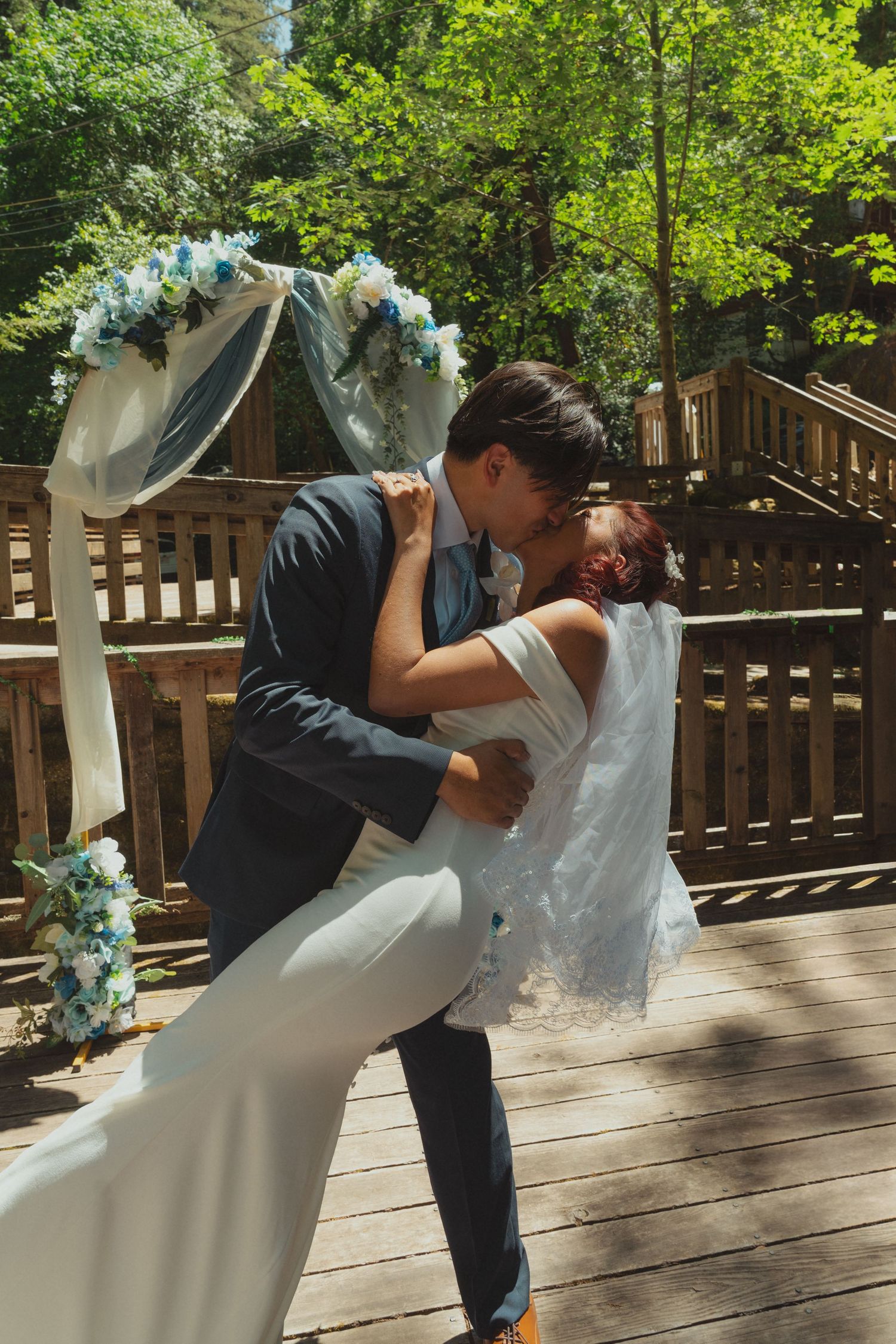 Bride and groom share a romantic dip kiss under a floral arch on a wooden deck surrounded by lush green trees.