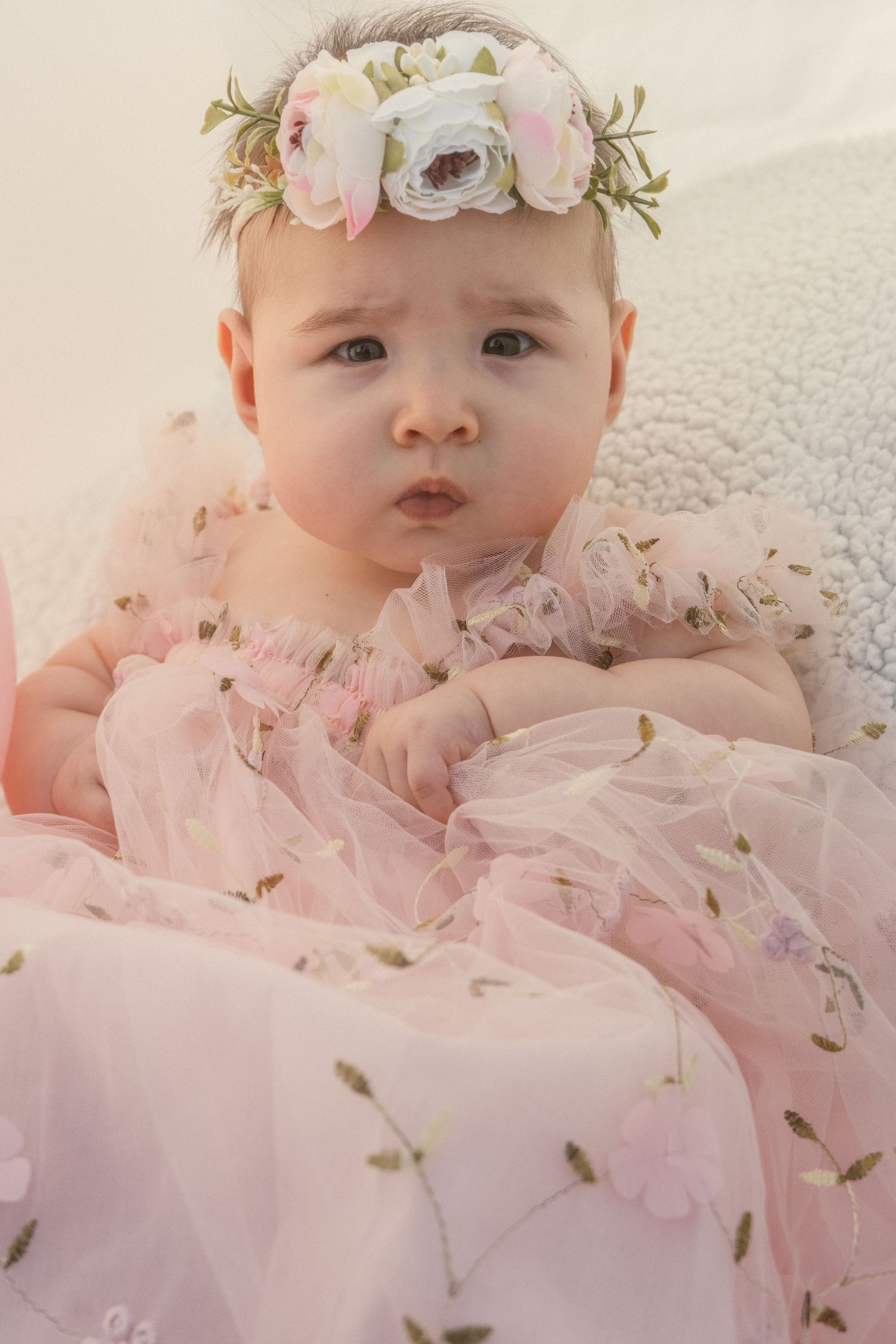 Baby in pink tulle dress with floral crown lying on cream textured blanket.