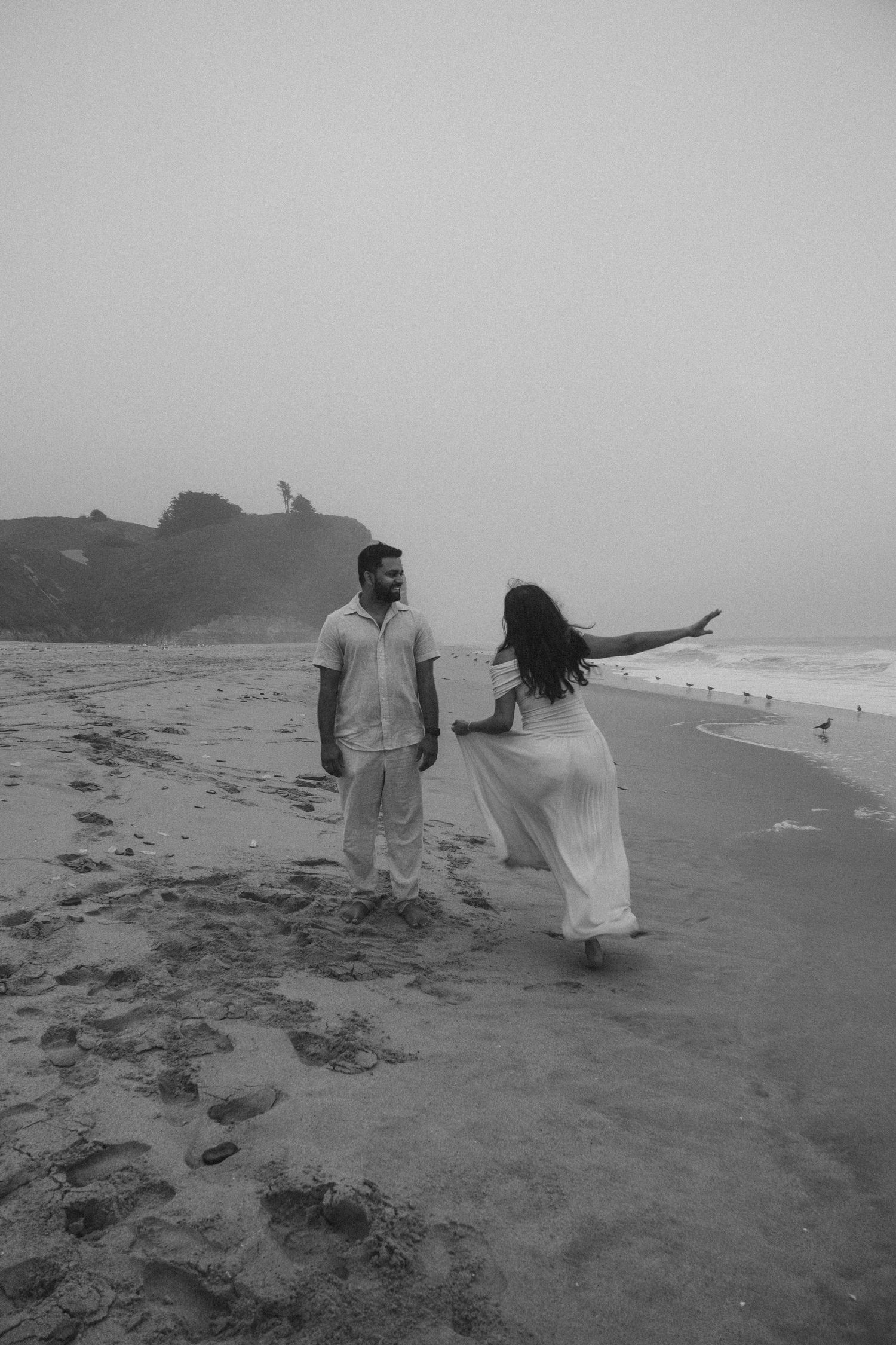A couple in white clothing walks along a misty beach with dramatic cliffs in the background.