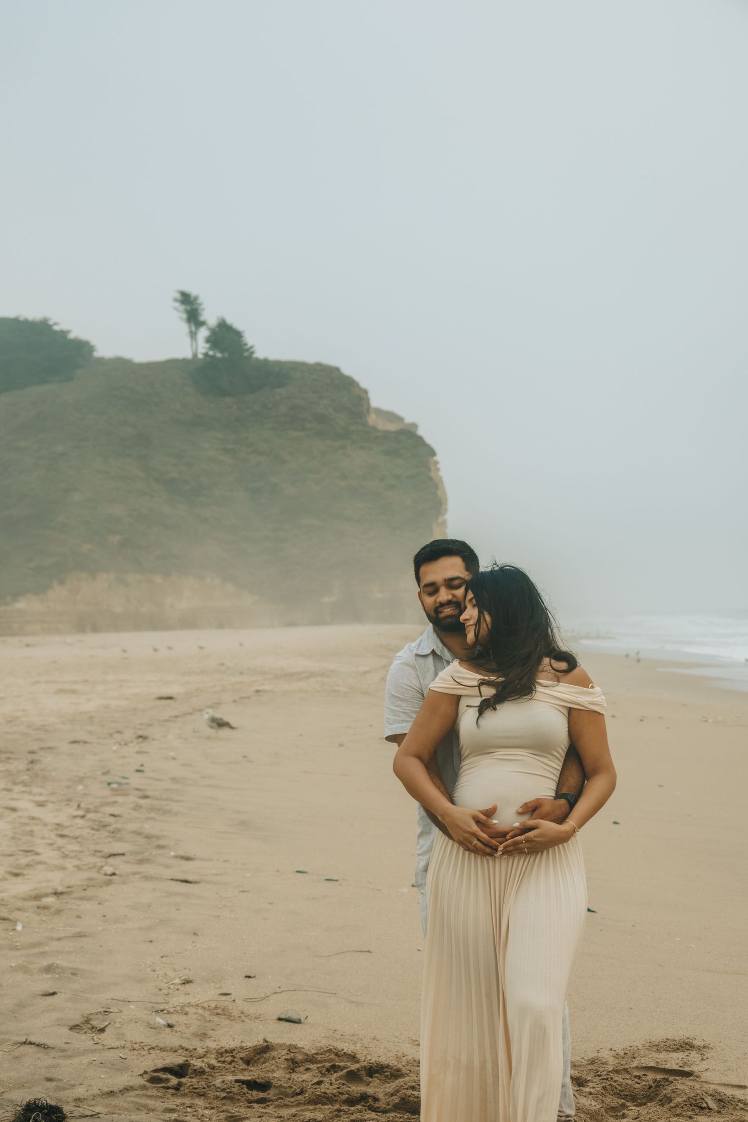 Couple shares intimate moment on beach with coastal cliffs in background.