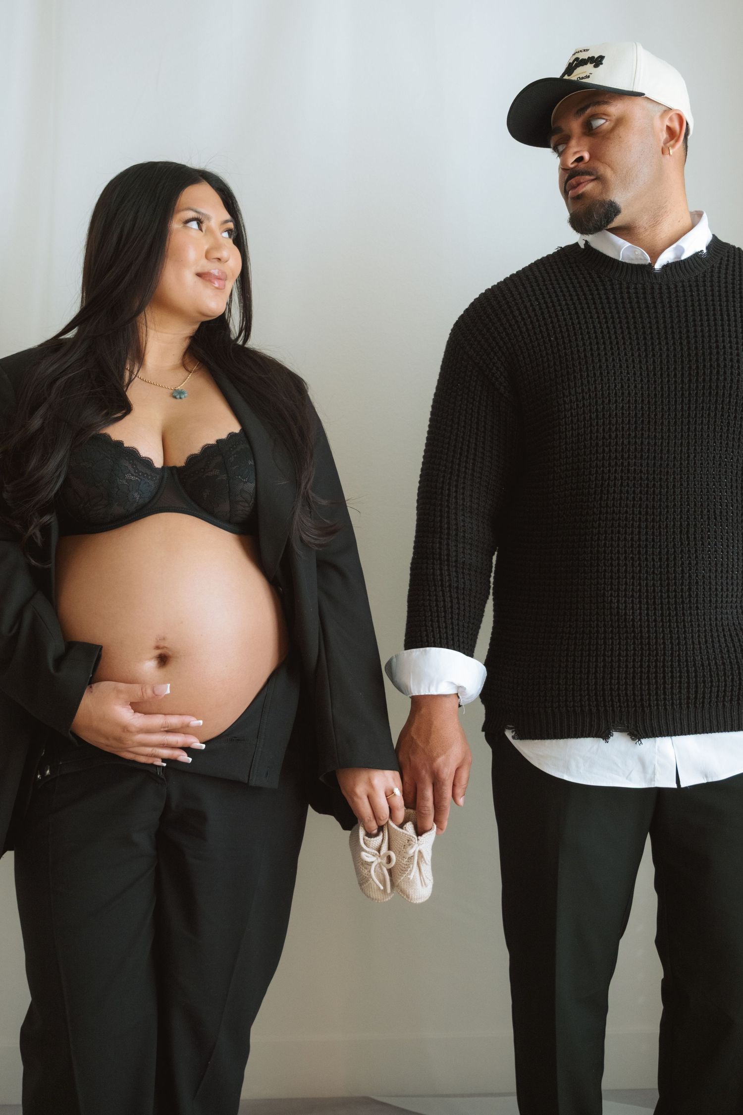 Pregnant couple in black outfits holding tiny baby shoes, expecting parents posing for maternity photo shoot.