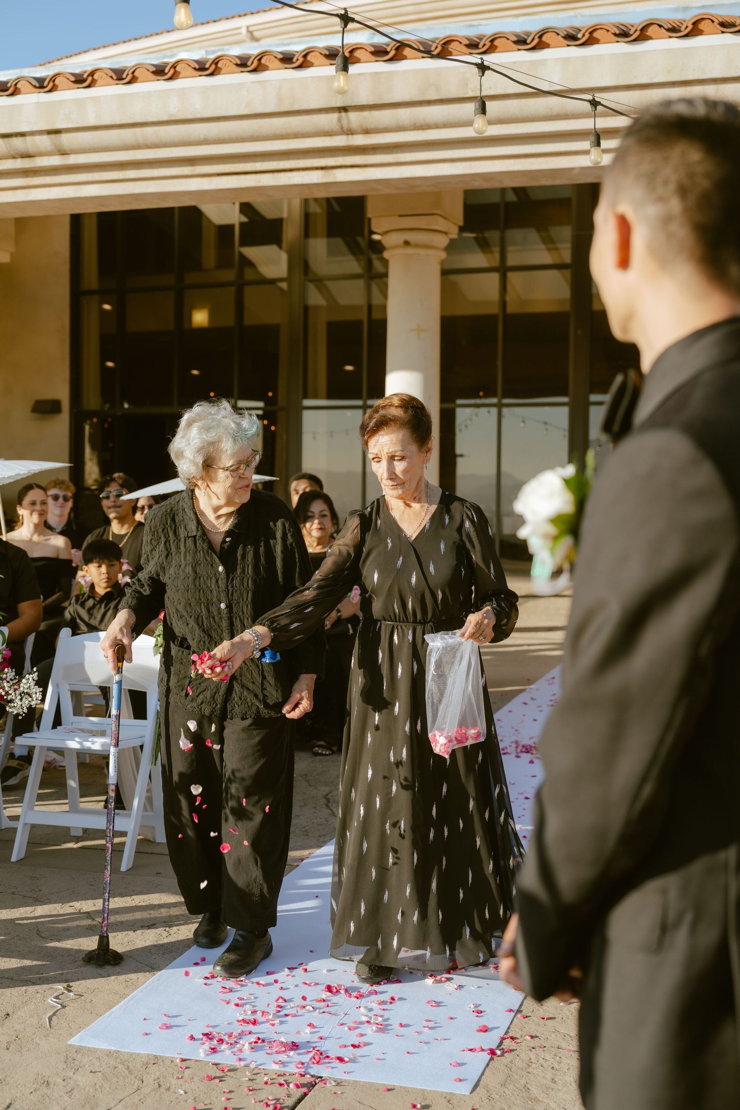 Two elderly flower girls scatter pink petals down wedding aisle at outdoor ceremony.