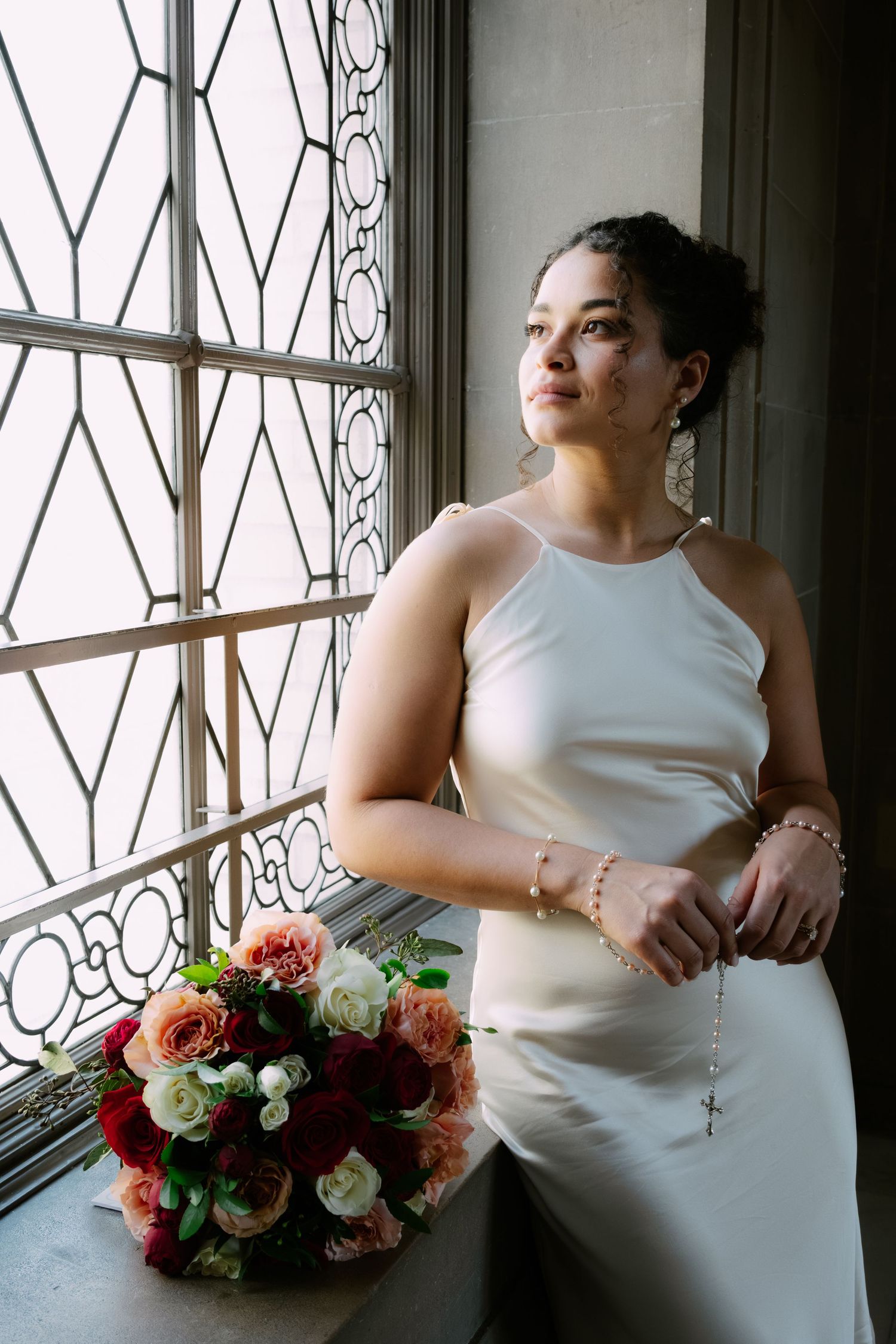 Bride in white halter gown holds rosary beads by ornate window with red and white floral bouquet nearby.