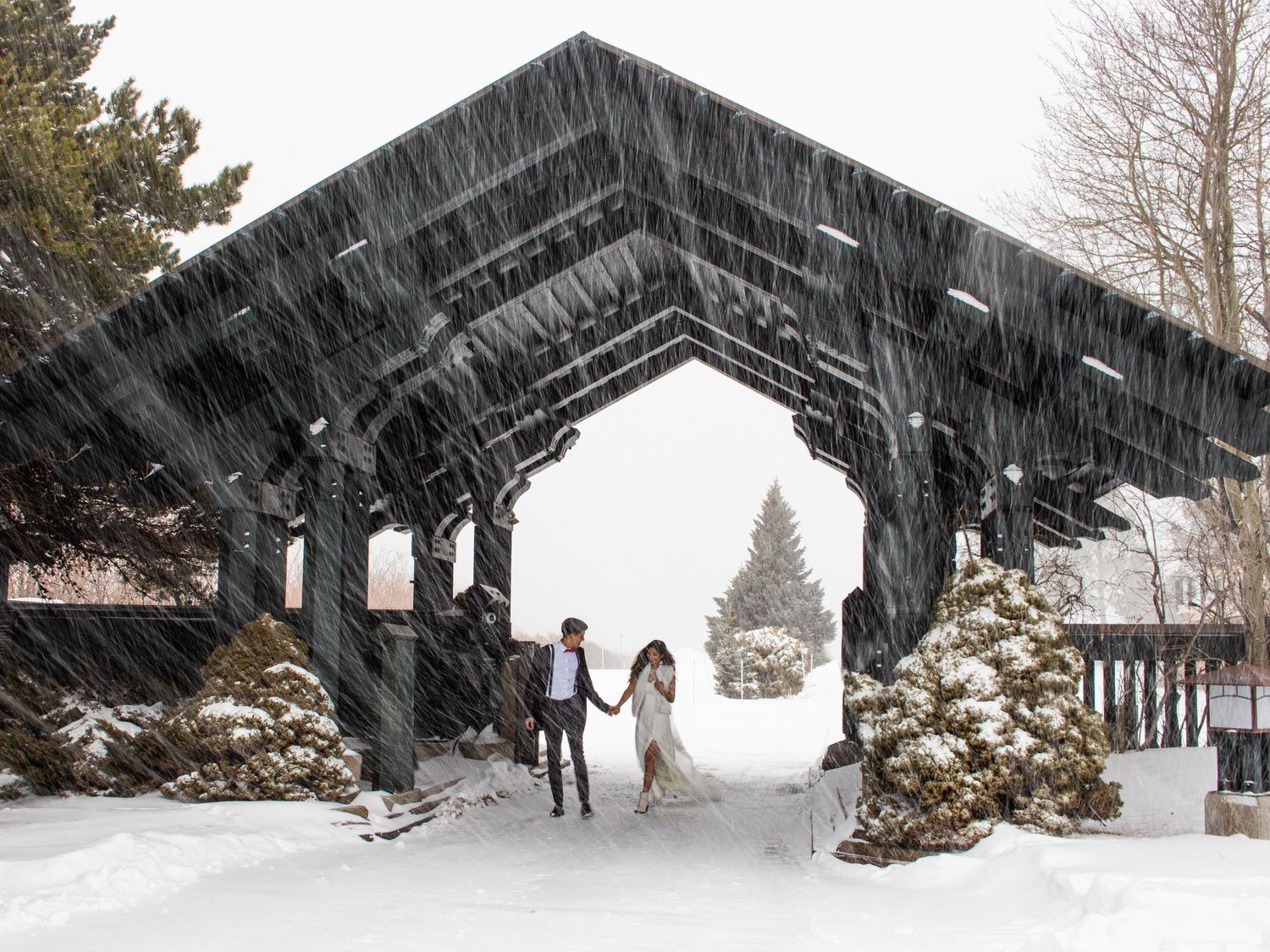 Winter wedding scene with couple walking through snow-covered wooden archway.