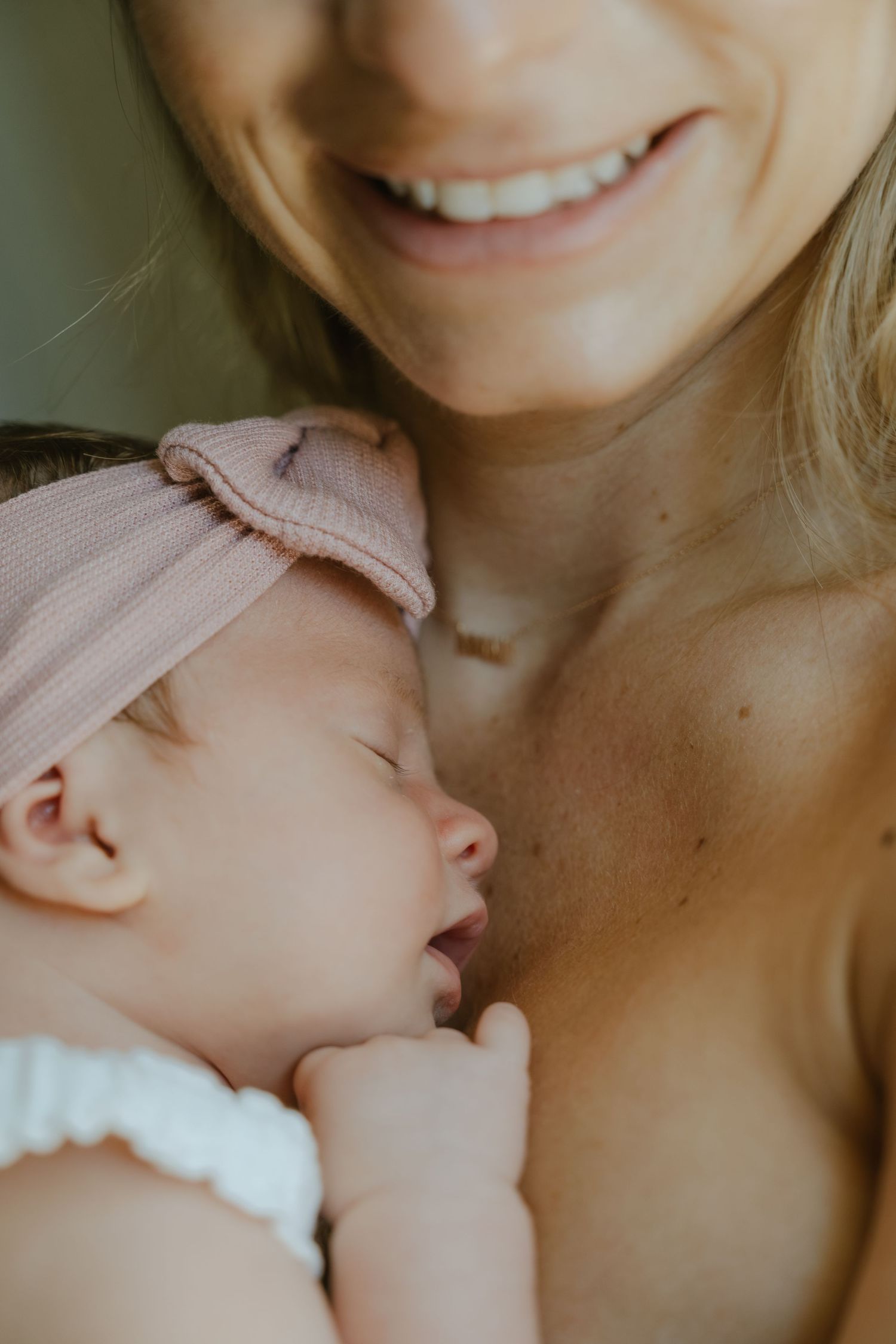 Smiling mother holding her sleeping newborn baby wearing a pink bow headband and white outfit close to her chest.