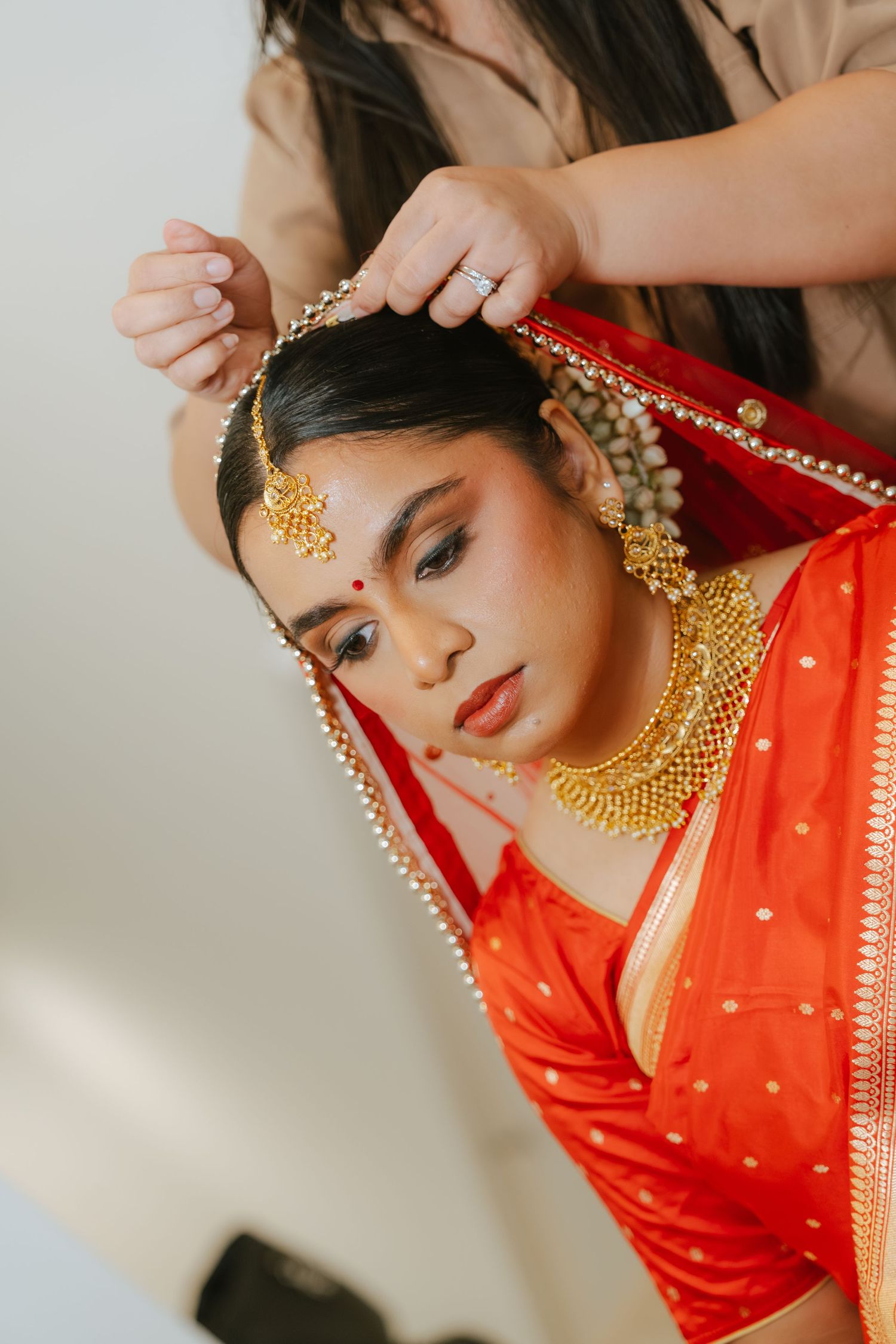 Indian bride in red saree and gold jewelry gets her dupatta arranged during bridal preparation.