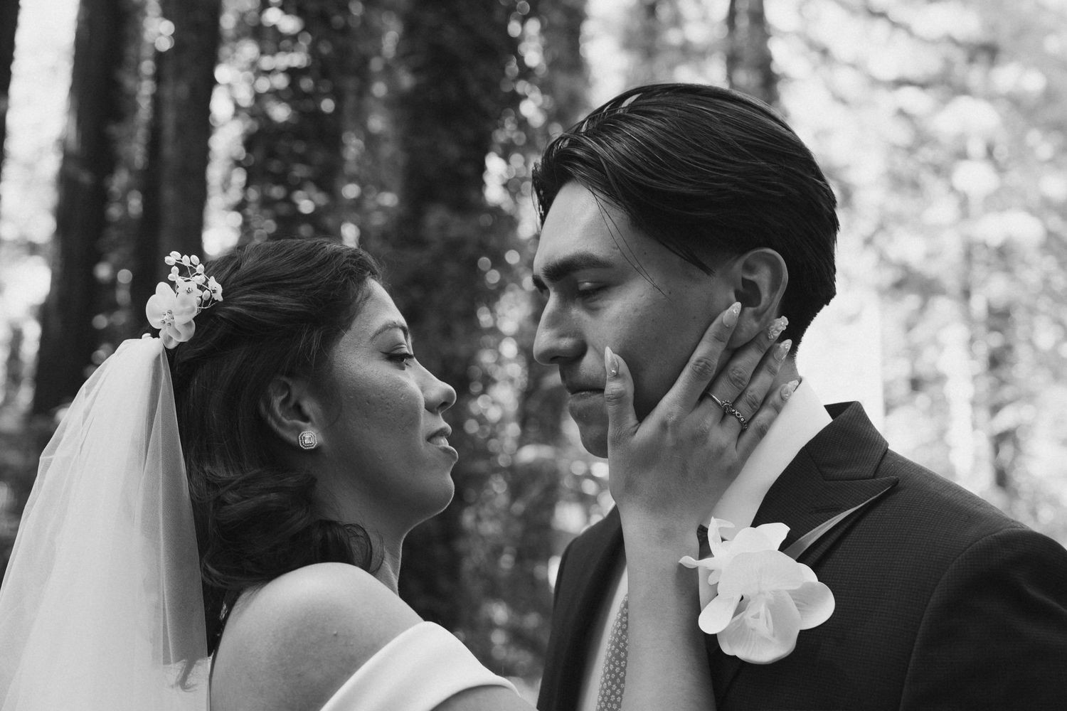 Black and white photo of bride tenderly cradling grooms face as they gaze at each other in a forest setting.
