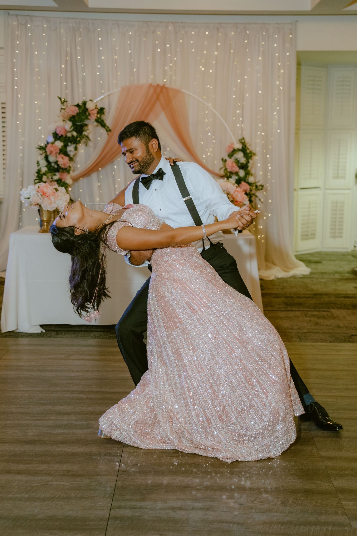 Couple shares first dance dip in sparkly pink gown at elegant wedding reception with floral arch backdrop.