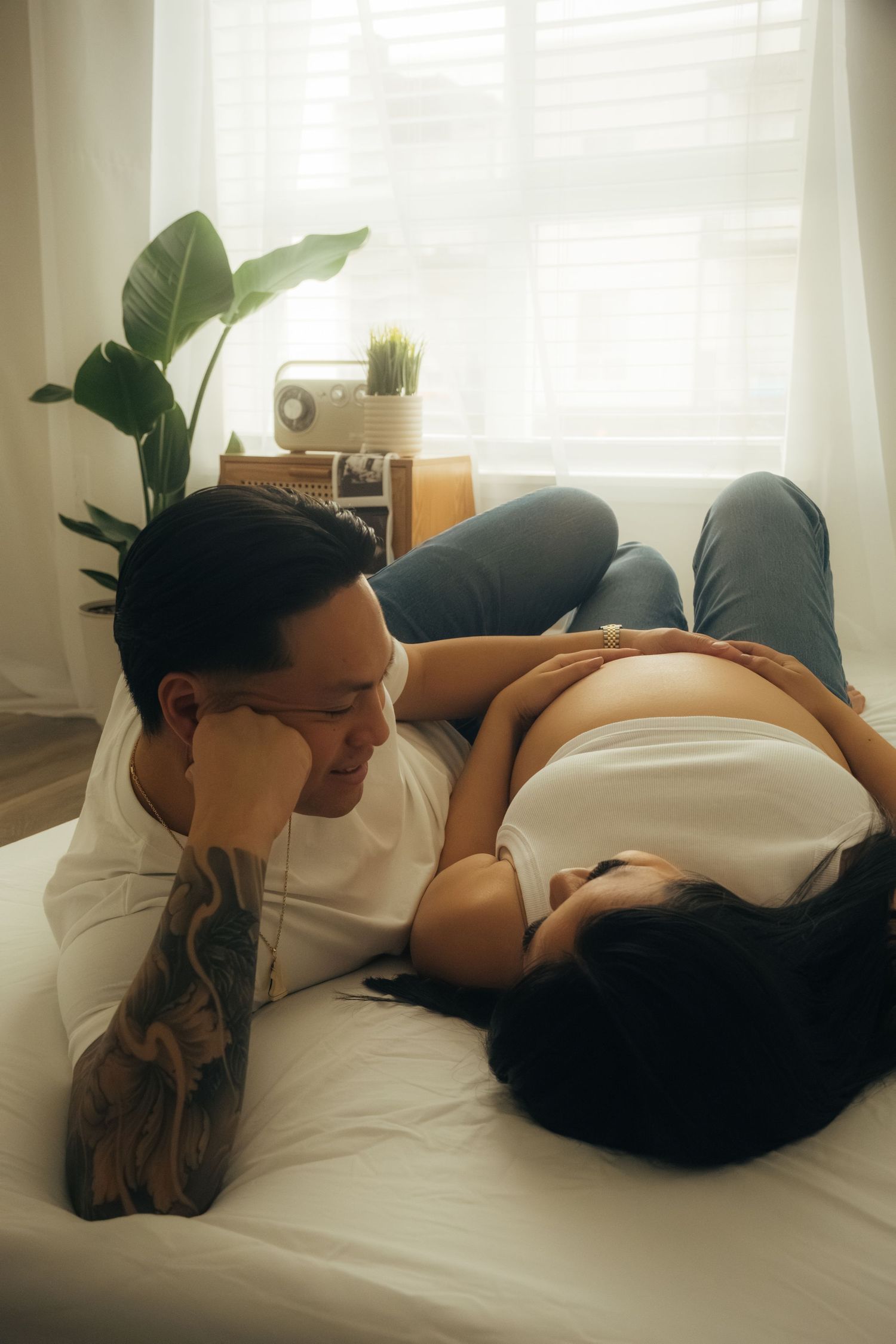 Relaxed maternity moment captured on white bed with houseplant in background.