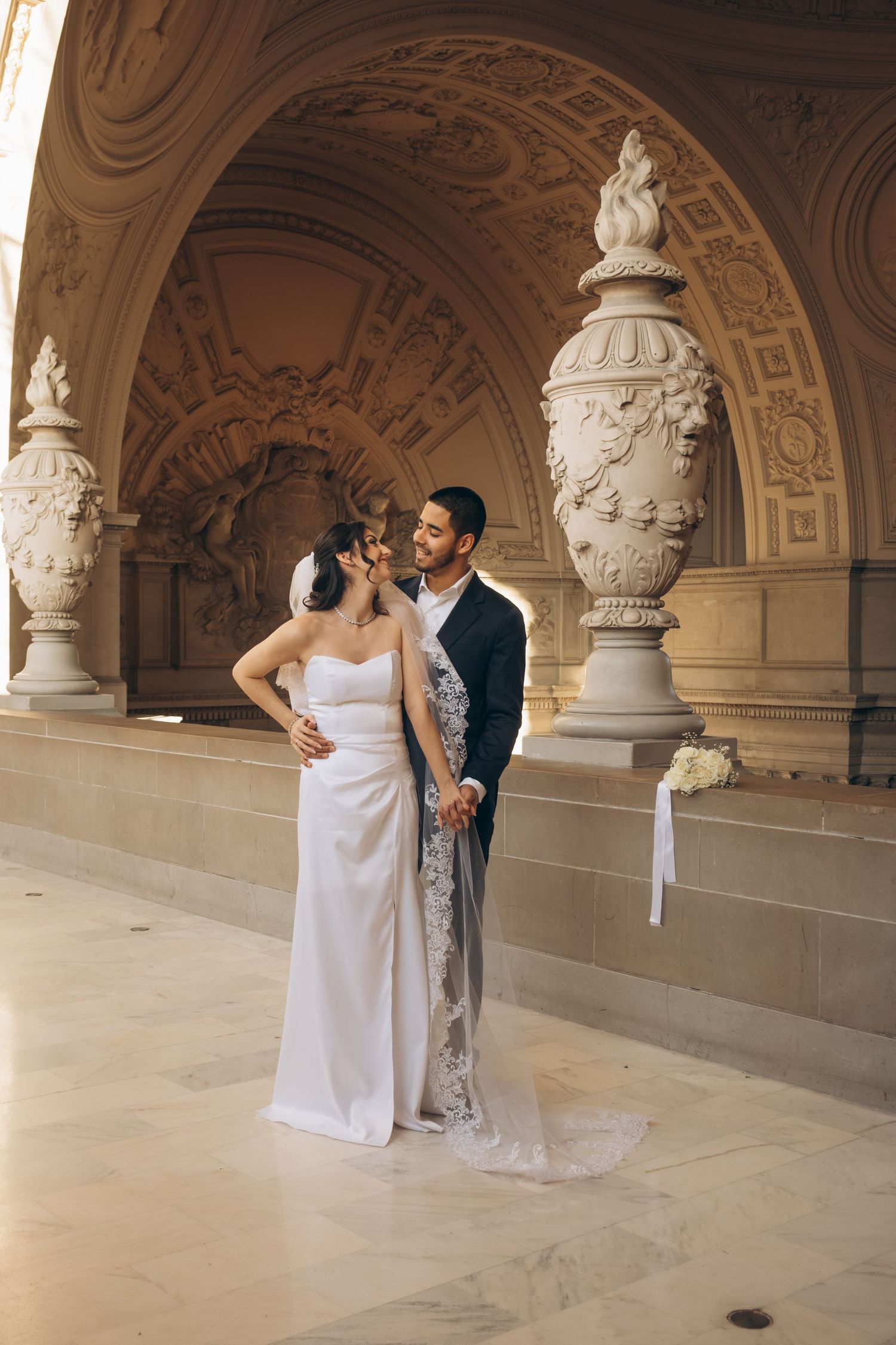 Wedding couple poses in an elegant hallway with classical sculptures and architectural details.