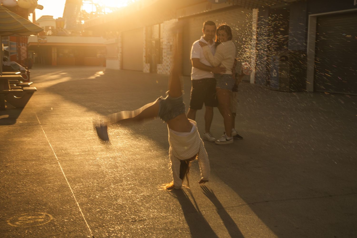 Child playing on sidewalk during sunset while adults watch in background.