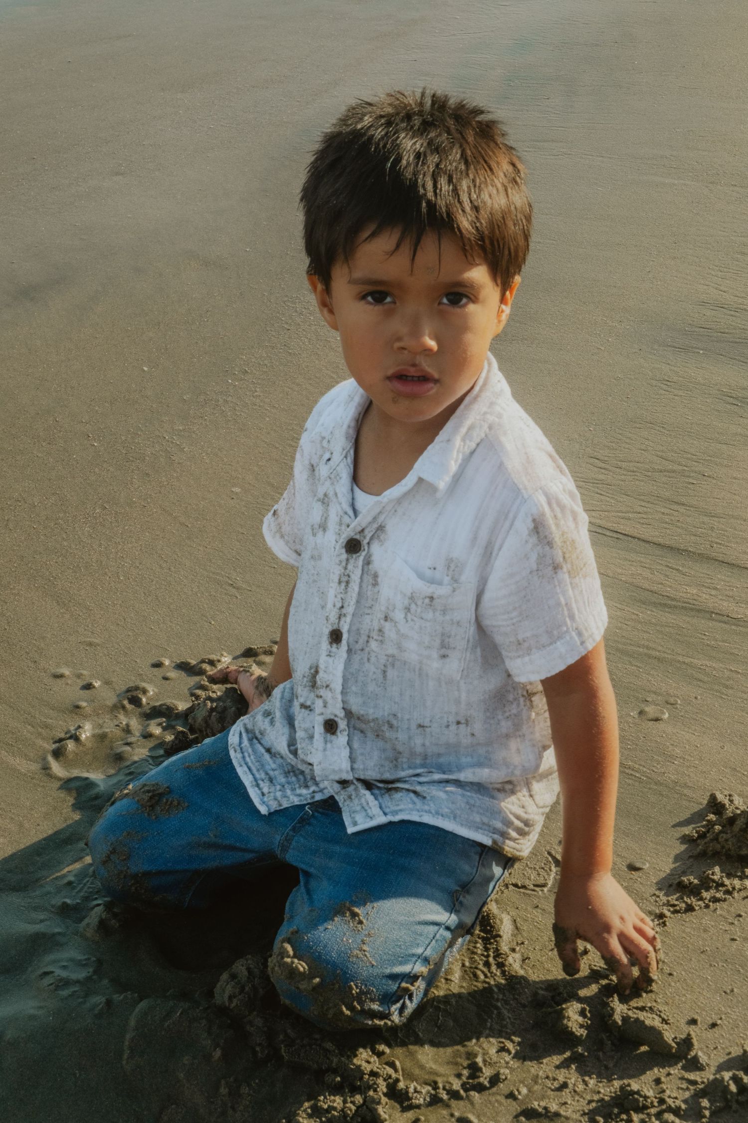 A small child in white shirt and jeans sits playing in the wet sand at the beach.