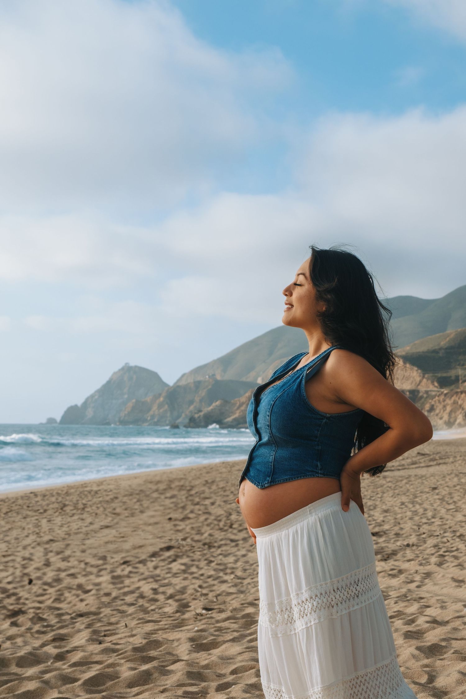 A person in denim and white stands on a scenic beach with mountains in the background.