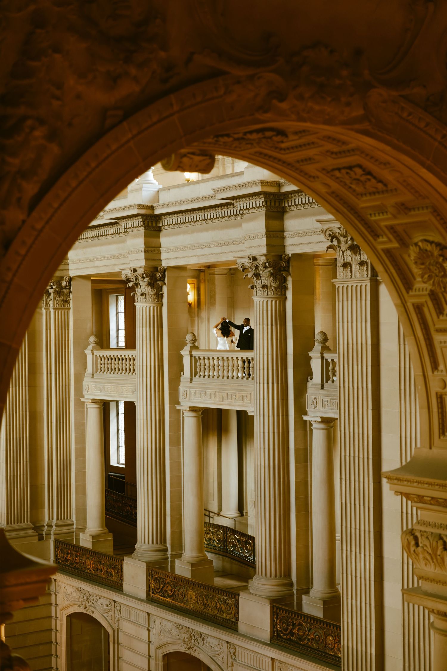 Couple dancing on ornate balcony framed by grand arch, surrounded by tall classical columns in elegant venue.