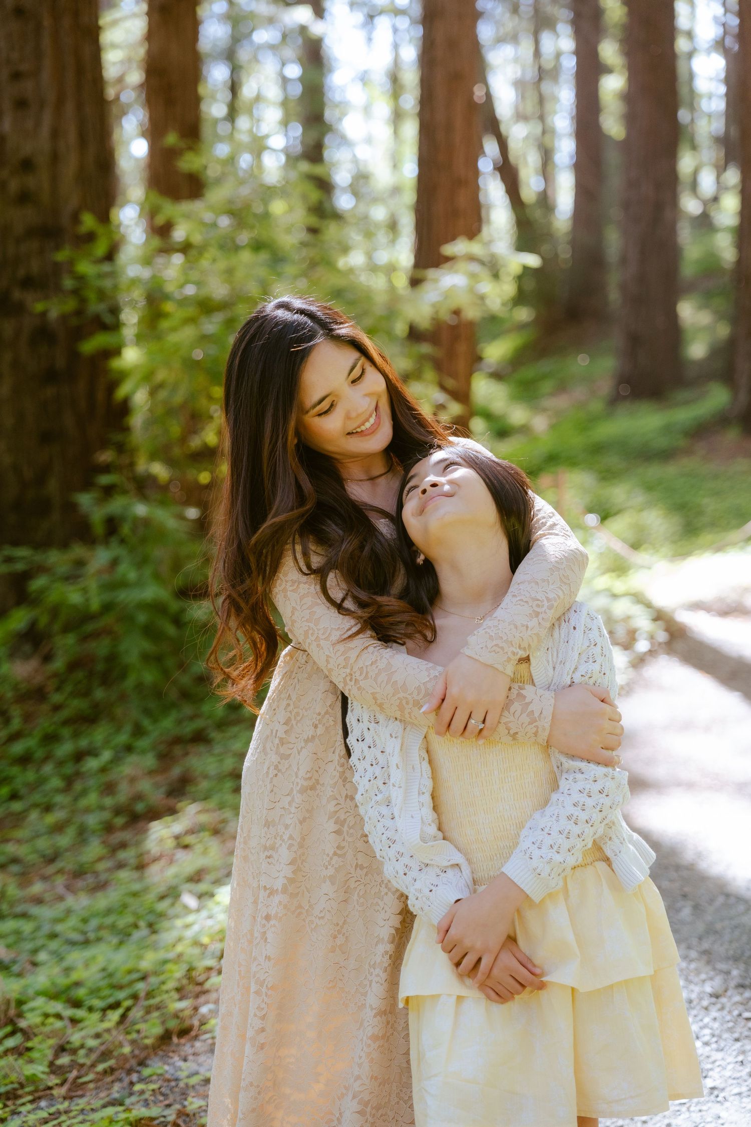 A mother and daughter share a warm embrace on a forest path, both wearing soft cream dresses among tall trees.