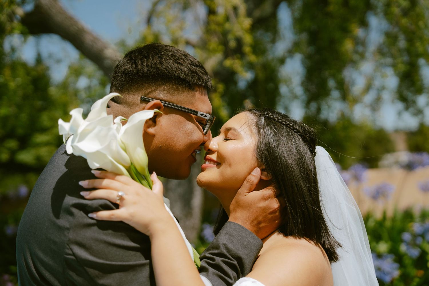 A bride and groom share a romantic moment, touching foreheads while holding white calla lilies outdoors on their wedding day.