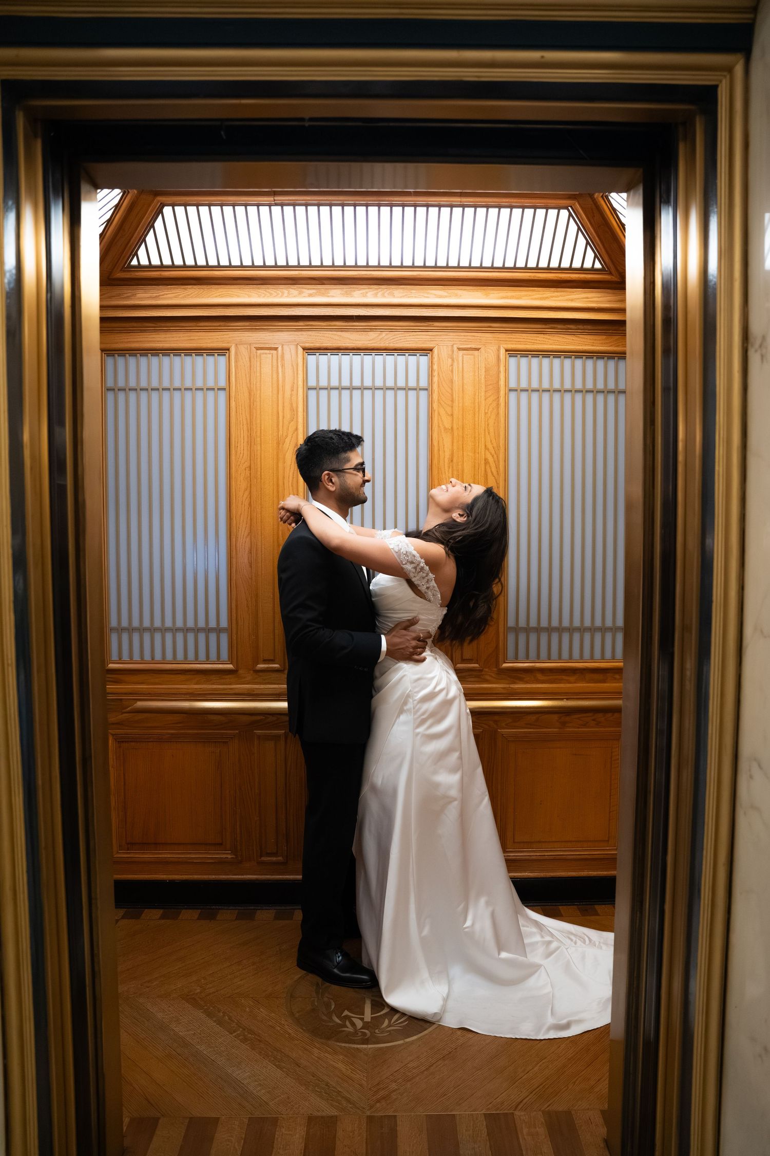 A bride and groom embrace romantically inside an elegant wooden elevator with frosted glass panels on their wedding day.