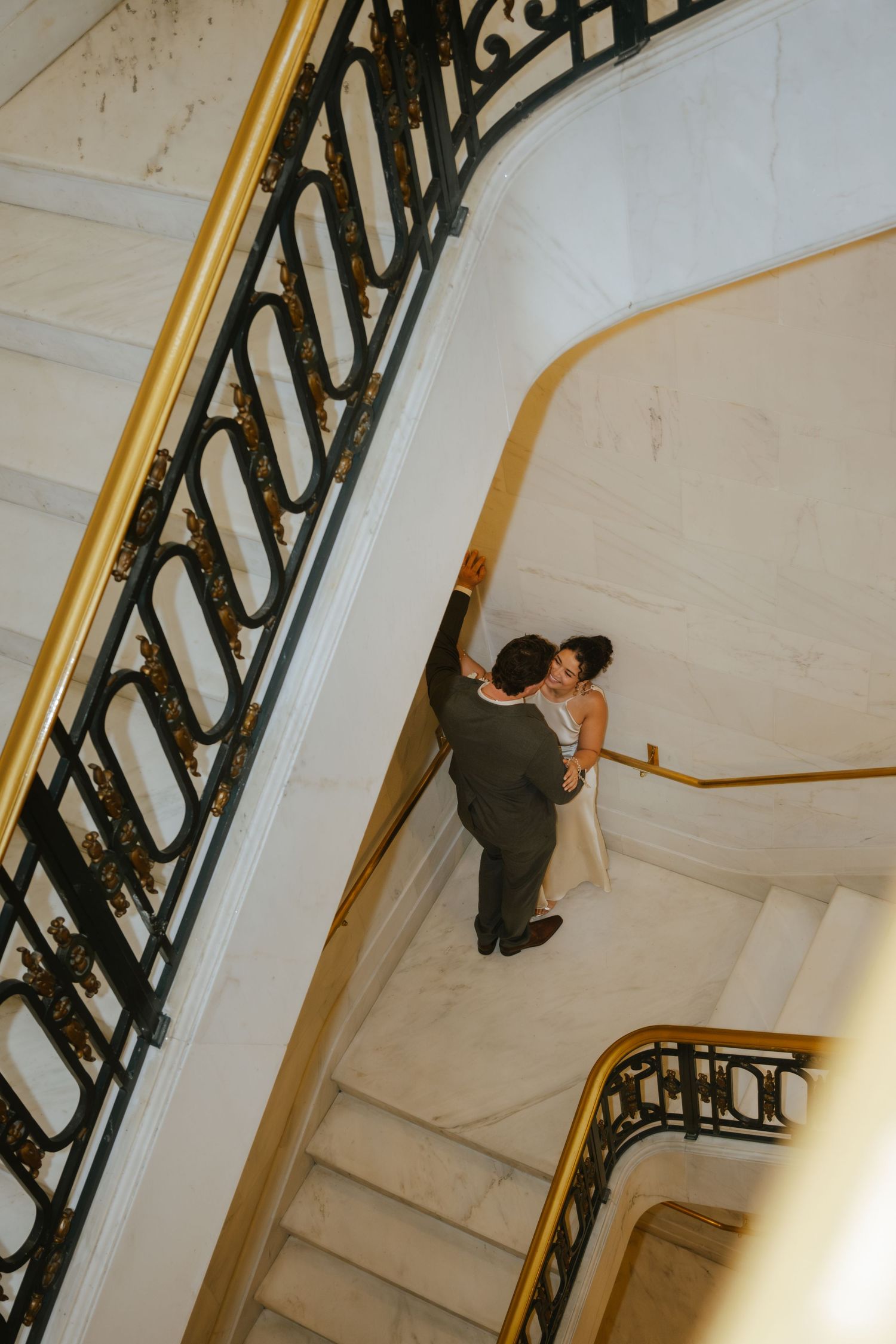 Aerial view of a wedding couple sharing a romantic moment on elegant marble stairs with ornate black and gold railings.