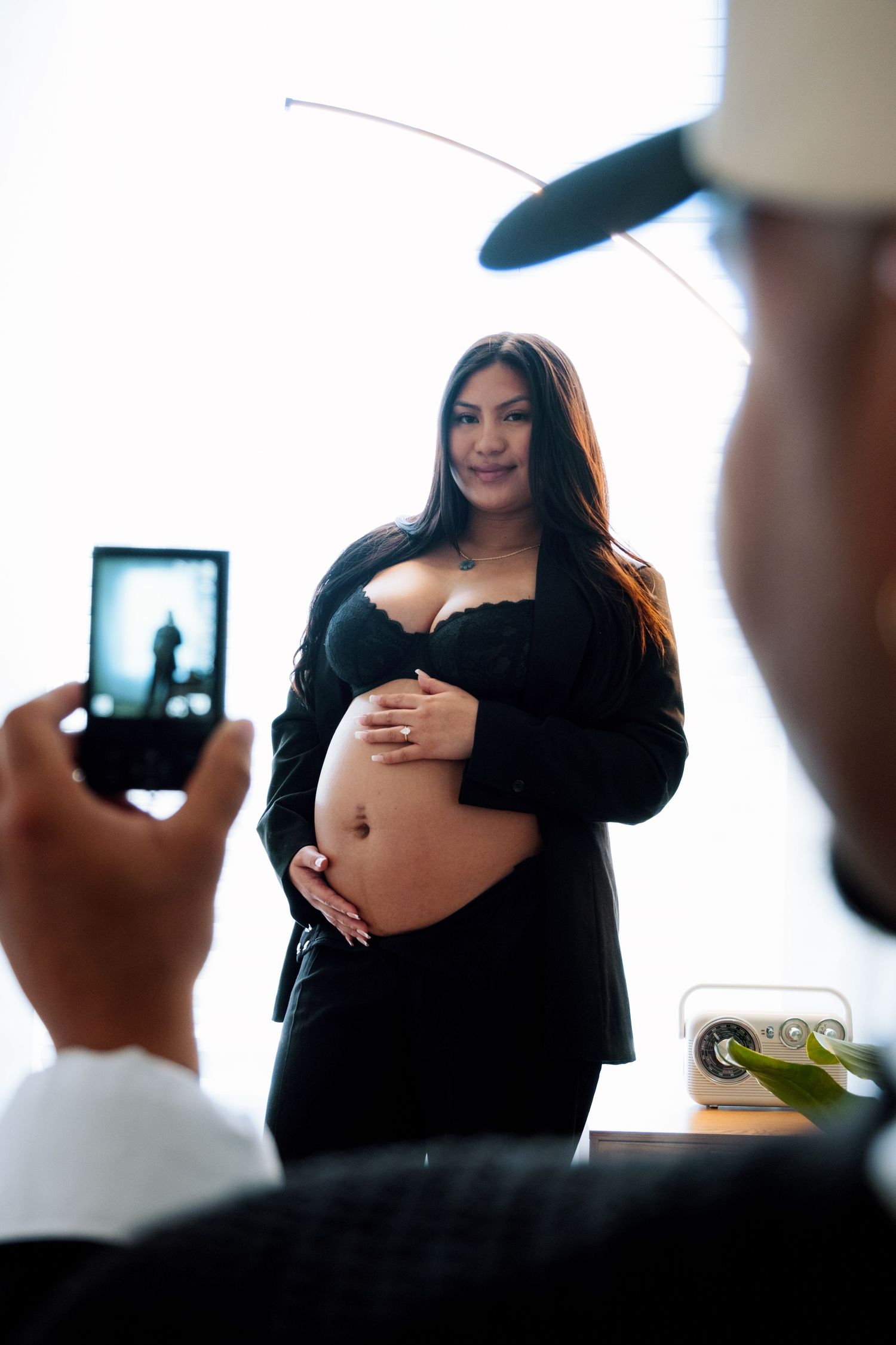 A maternity photoshoot in progress showing someone in black clothing posing while another person captures the moment on a phone.