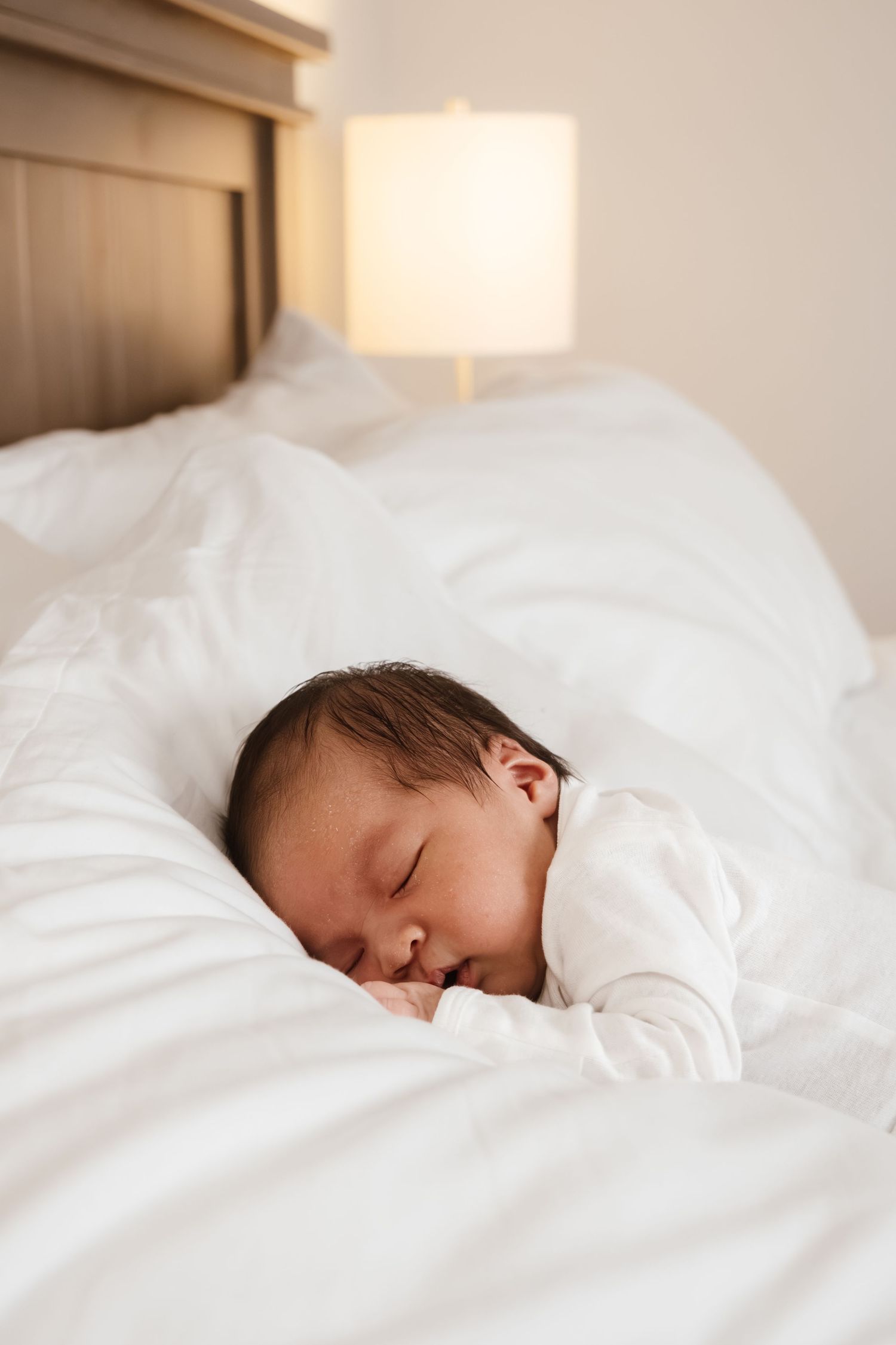 A sleeping baby peacefully rests in white bedding with soft natural light streaming across the scene.