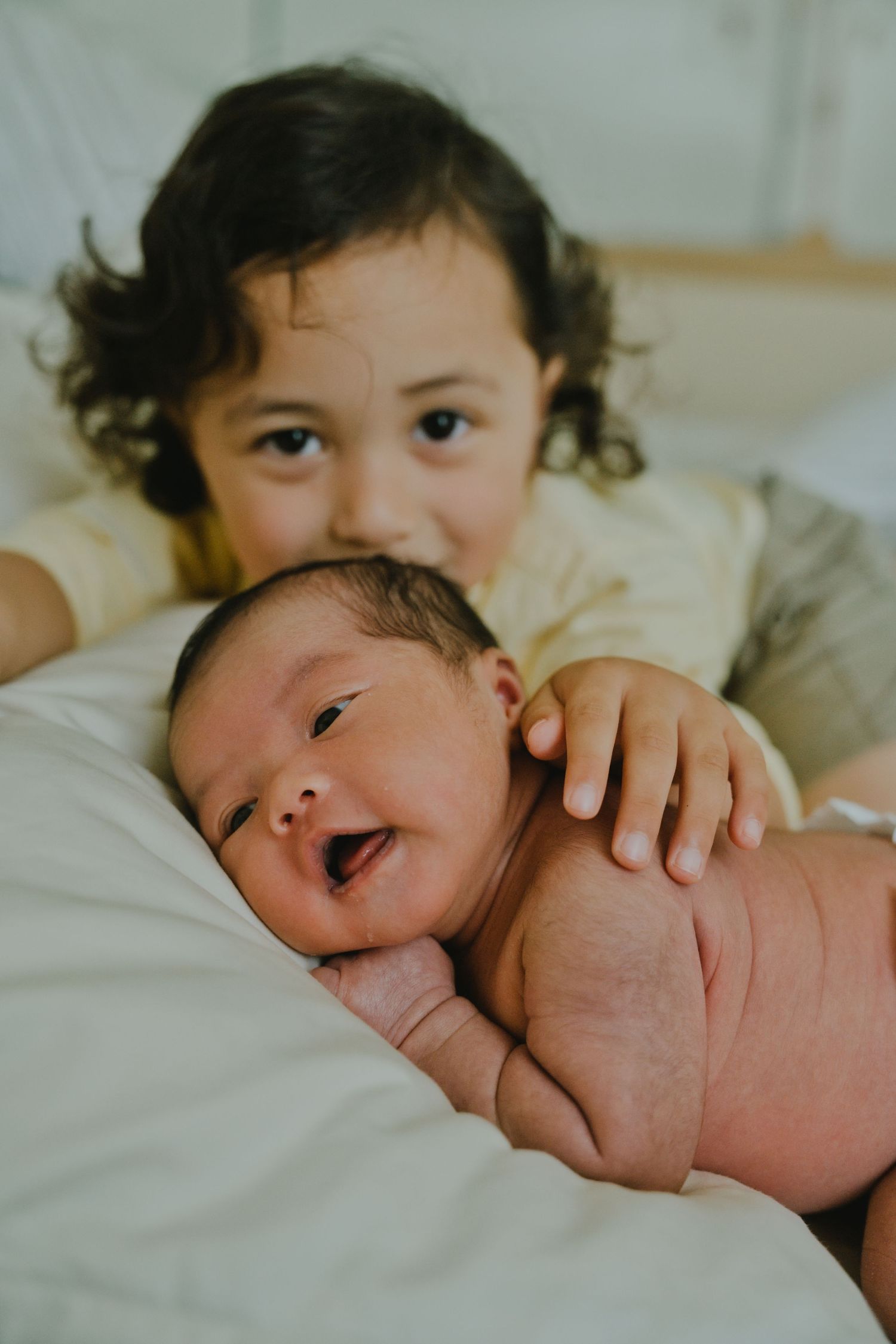 Young child smiling behind a laughing newborn baby lying on a white bed, siblings bonding moment.