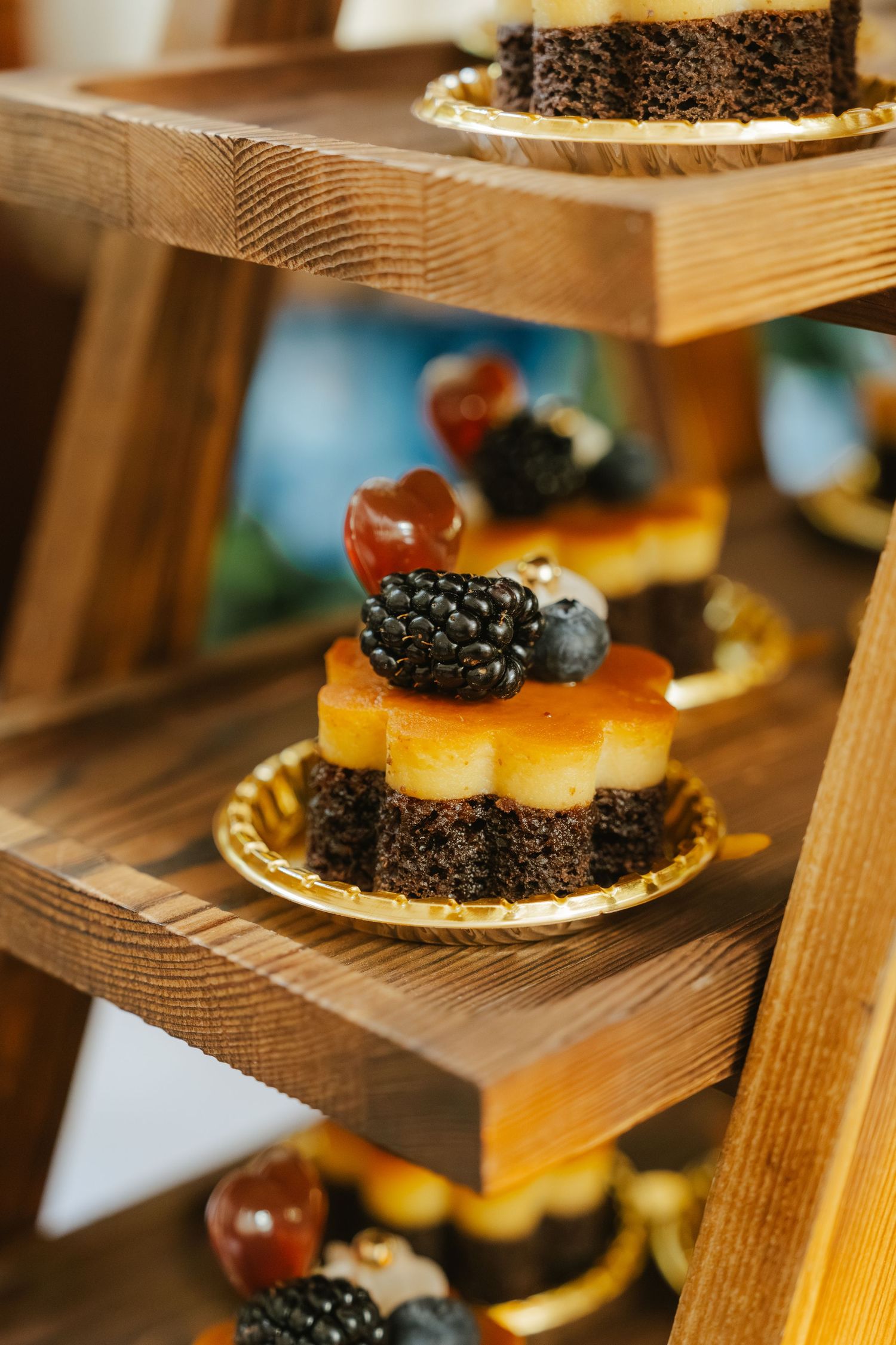 Elegant wooden tiered dessert stand displaying chocolate petit fours topped with fresh blackberries, blueberries, and caramel glaze.