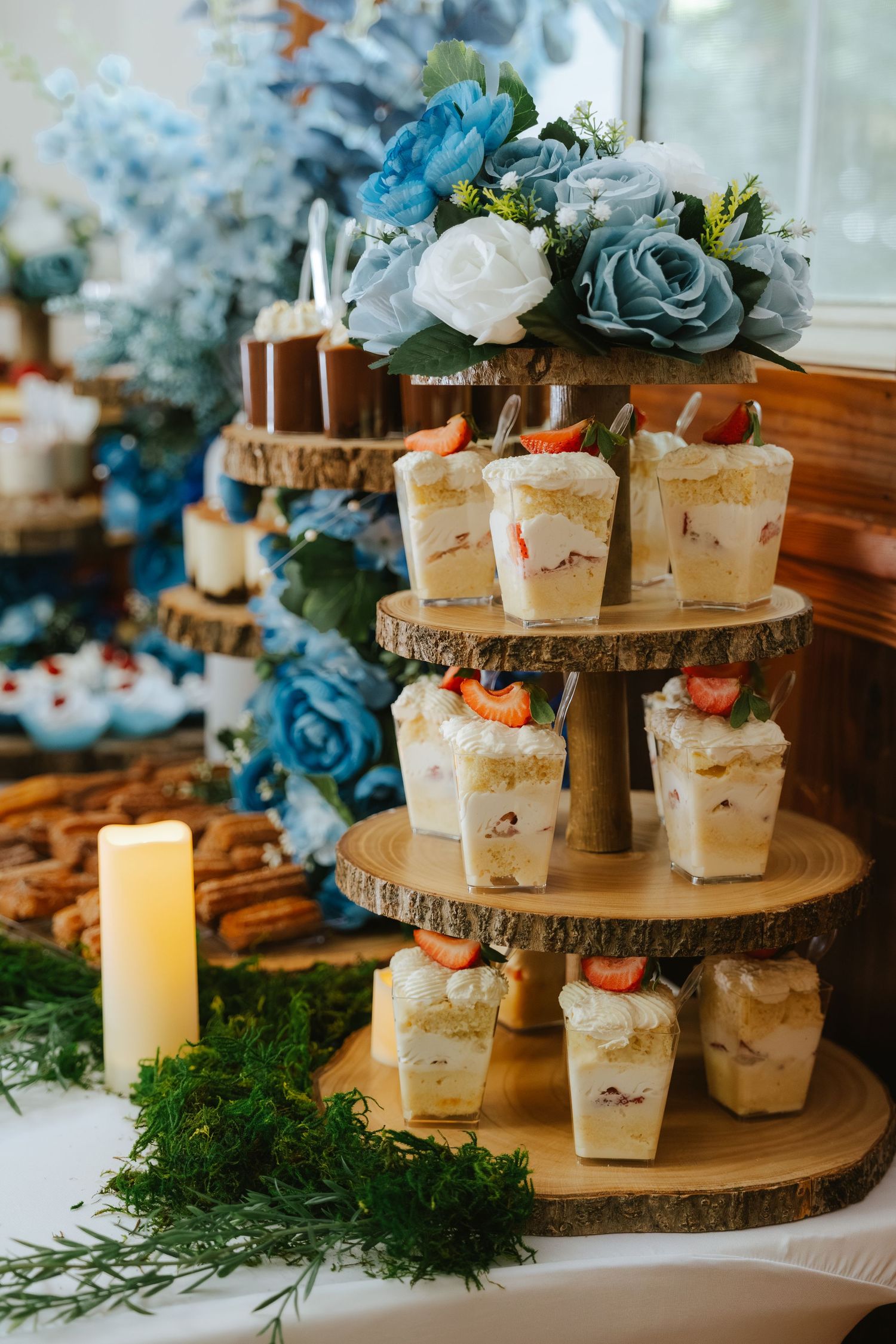 Rustic wooden tiered dessert stand with strawberry trifle cups, blue roses, and candles at an elegant wedding reception.